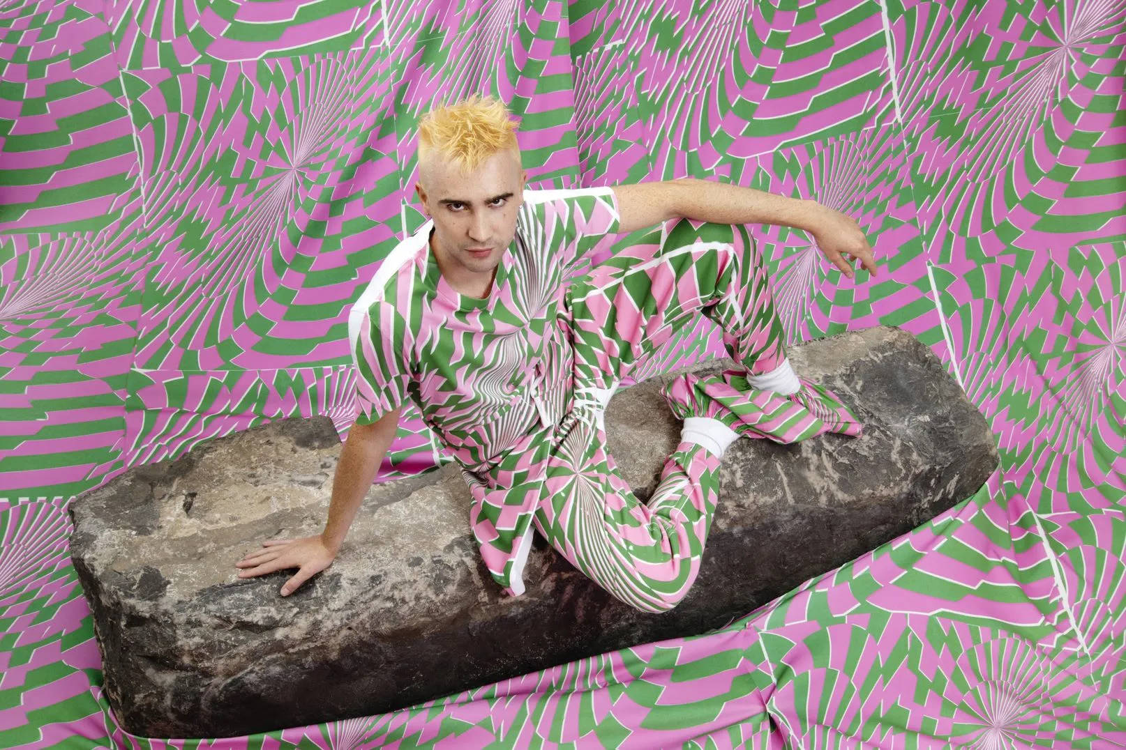 Stella McCartney x Ed Curtis 壁紙 — 一個金髮的人自信地坐在一塊大石頭上，背景是充滿活力的粉紅色和綠色幾何圖案。