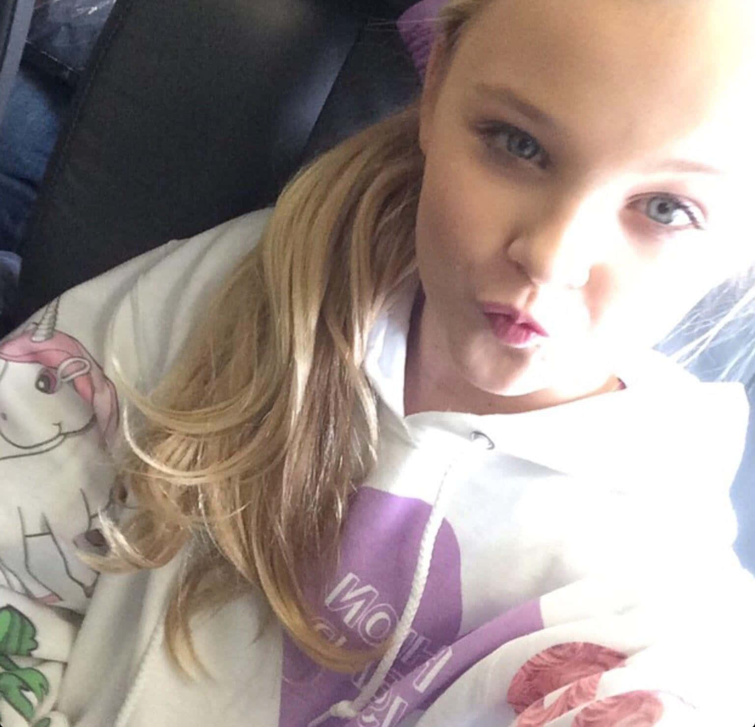 jojo siwa 桌布 壁紙 — 一個金髮藍眼的小女孩在汽車座椅上穿著獨角獸連帽衫，做出頑皮的表情。