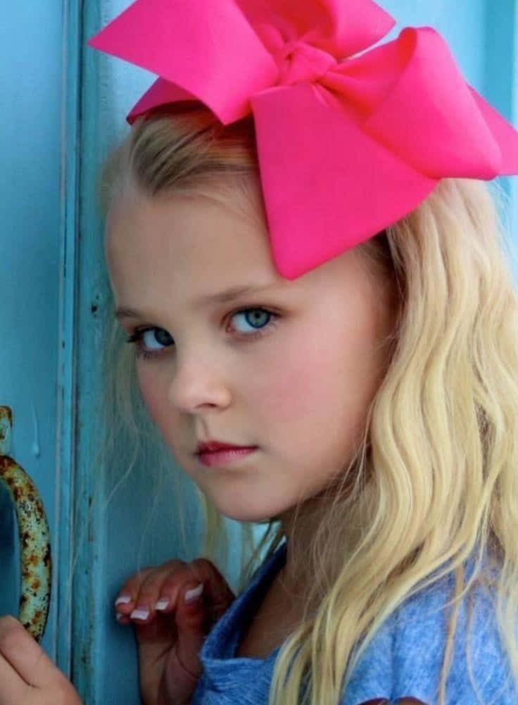 jojo siwa 桌布 壁紙 — 一個金髮藍眼的小女孩戴著一個大粉紅色蝴蝶結，靠在一扇風化的藍色門上。