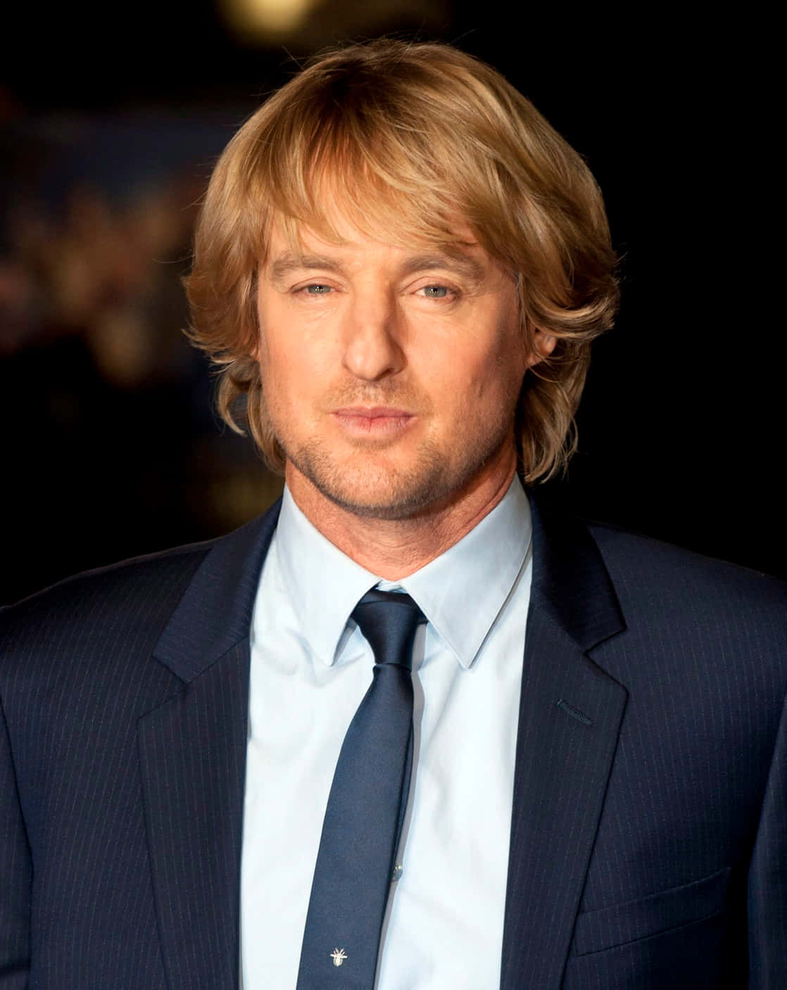 Owen Wilson 桌布 壁紙 — 一個金髮鬍鬚男士的特寫肖像，穿著深色細條紋西裝和領帶，背景模糊。