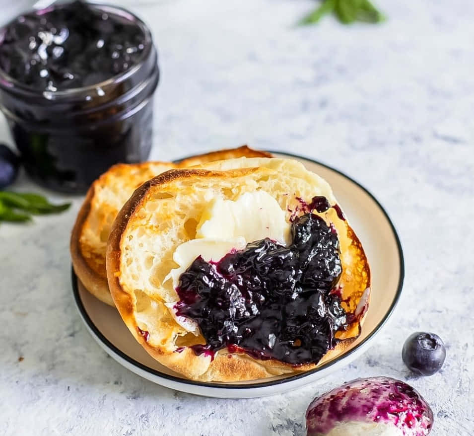 Blueberry Jam 桌布 壁紙 — 一個金黃色的烤麵包卷切開，配上奶油狀的牛油和濃郁的深色藍莓果醬，放在一個有斑點的碟子上。