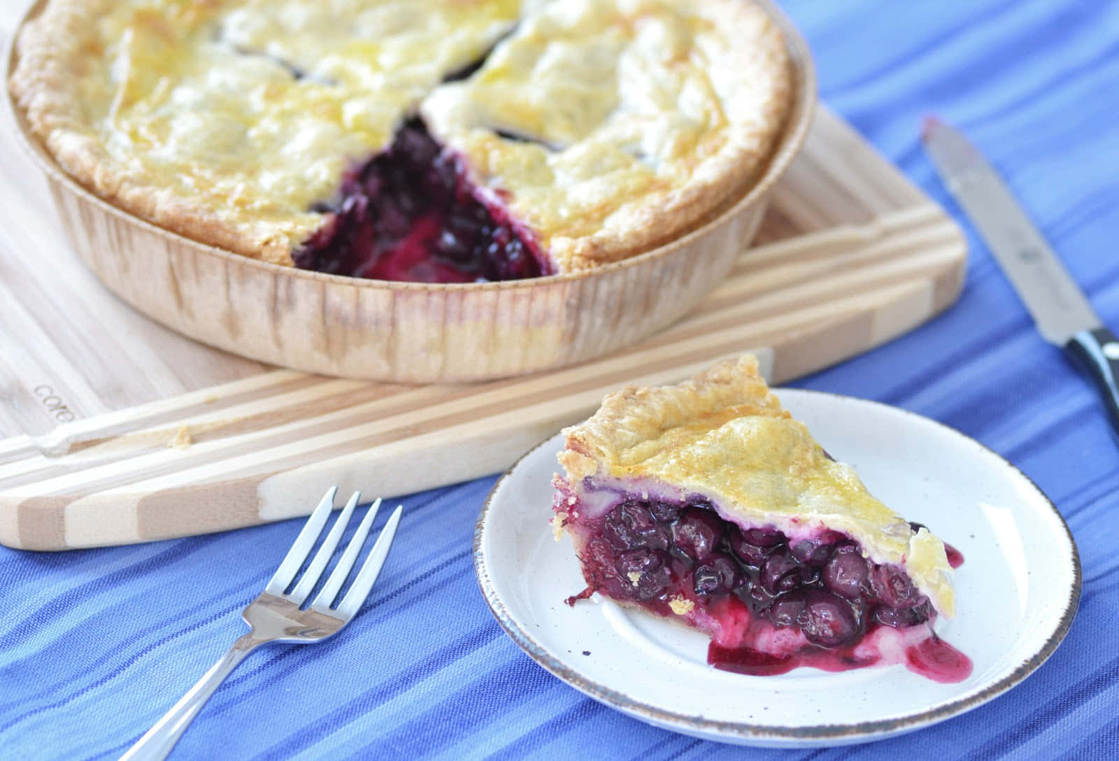 Blueberry Pie 桌布 壁紙 — 一個金黃色的藍莓批，帶有酥脆的外皮，放在木盤中，旁邊是一塊放在碟上的切片，置於藍色條紋布上。