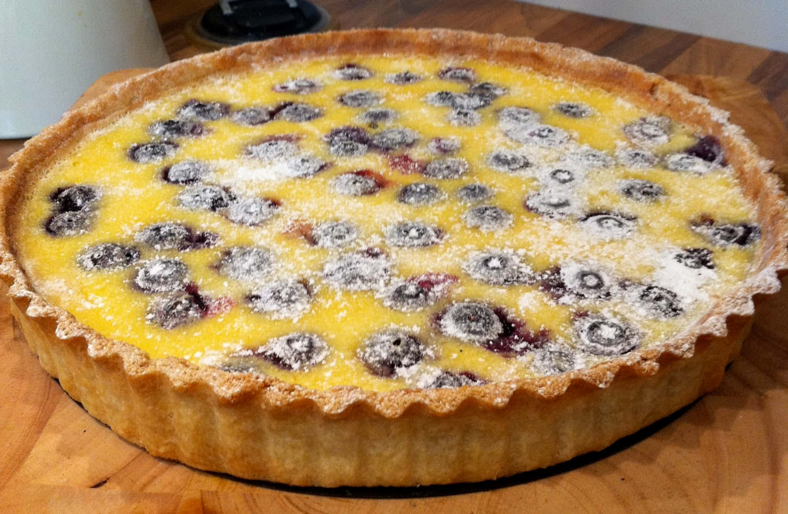 Blueberries Tart 桌布 壁紙 — 一個金黃色的藍莓撻撒上糖粉，放在木製砧板上。