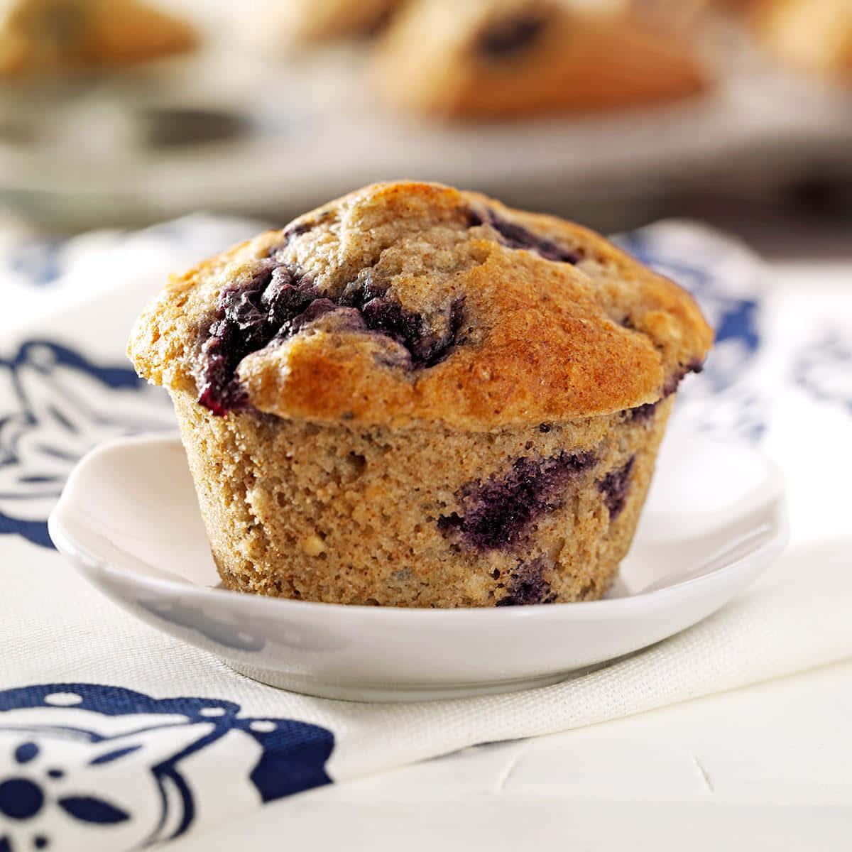 Blueberry Muffins 桌布 壁紙 — 一個金黃色的藍莓鬆餅，上面有多汁的紫色莓果，放在一個白色碟子上，碟子下面是一塊藍白圖案的布。