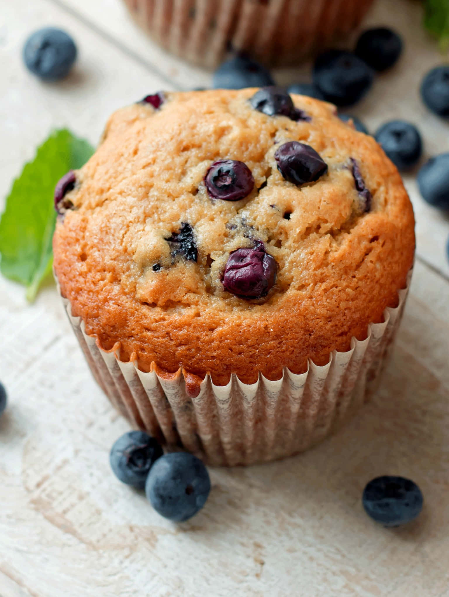 Blueberry Muffins 桌布 壁紙 — 一個金黃色的藍莓鬆餅，上面有飽滿的藍莓，放在質樸的木質表面上，周圍是新鮮藍莓和一片薄荷葉。