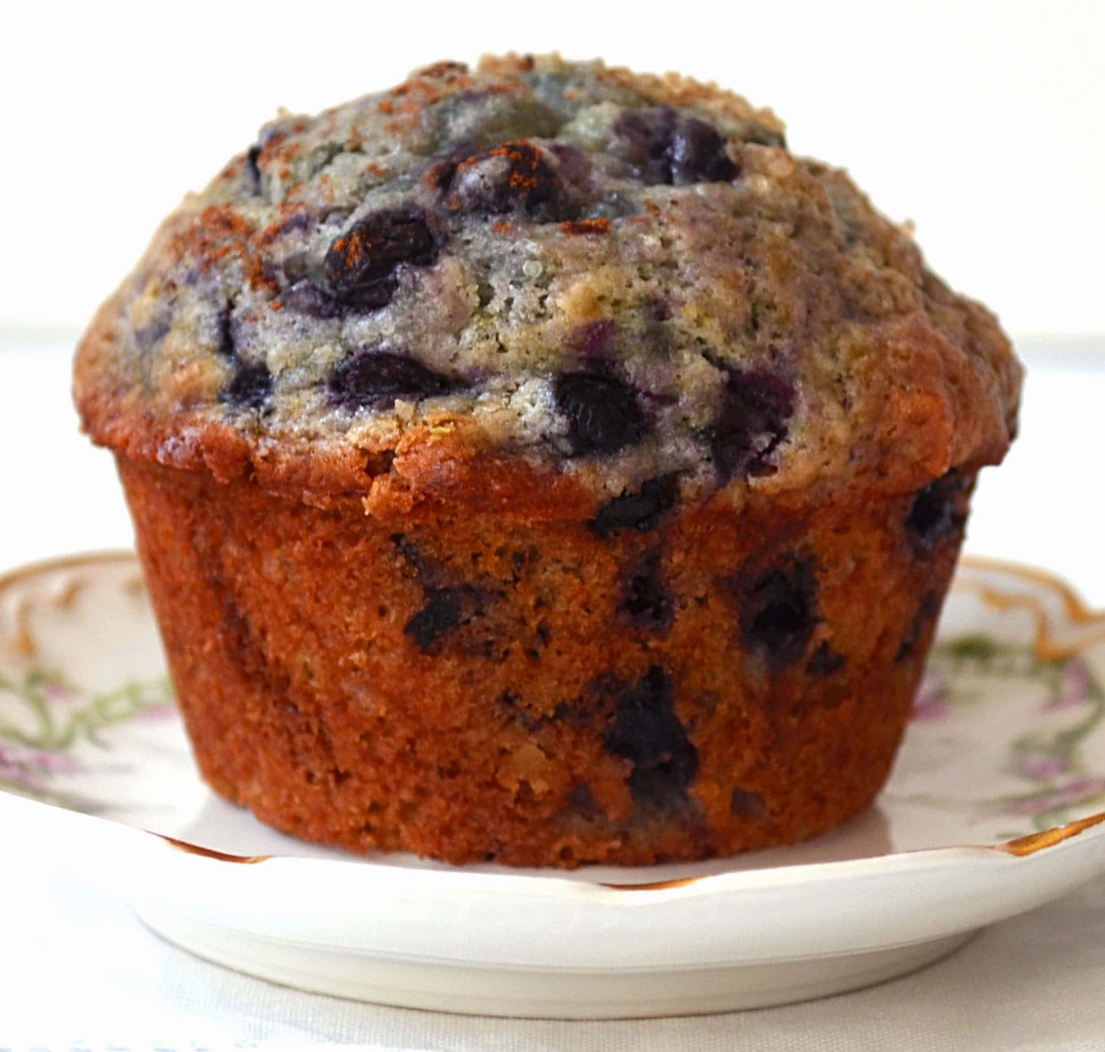 Blueberry Muffins 桌布 壁紙 — 一個金黃色的藍莓鬆餅，帶有爆裂的莓果，放在一個花卉圖案的碟子上，背景是明亮的白色。