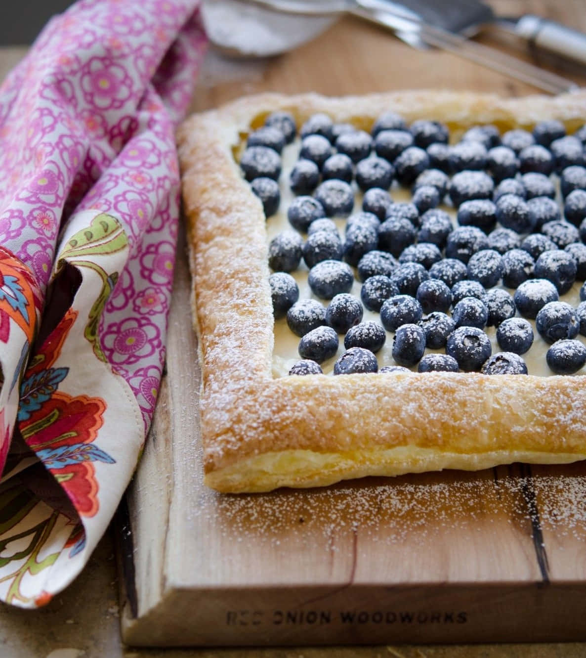Blueberries Tart 桌布 壁紙 — 一個金黃色的酥皮撻，上面鋪滿新鮮藍莓和糖粉，放在木板上，旁邊有一塊色彩繽紛的花布。