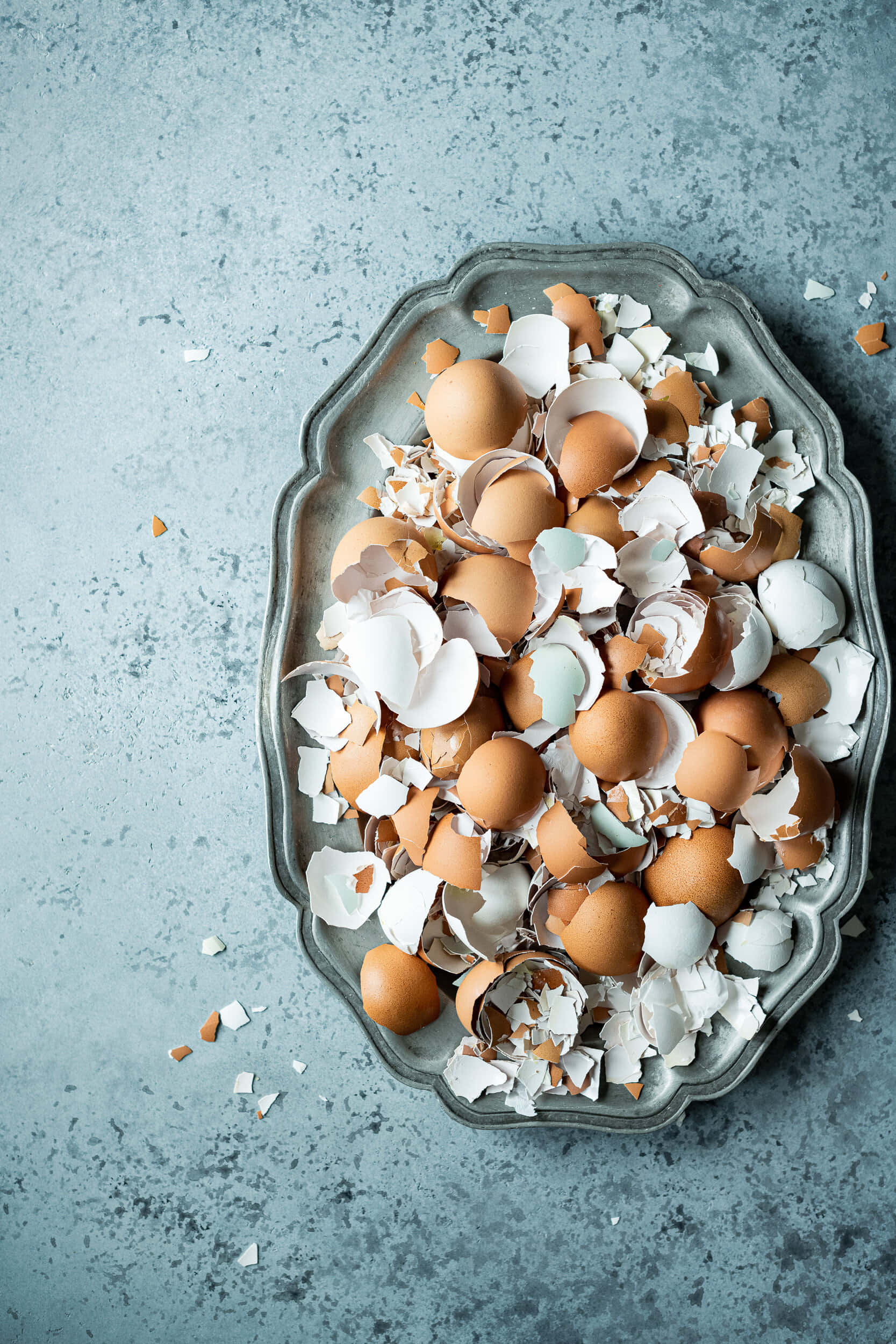 Eggshell 桌布 壁紙 — 一個銀托盤上堆滿了破碎的棕色和白色蛋殼，放在有紋理的藍灰色表面上。