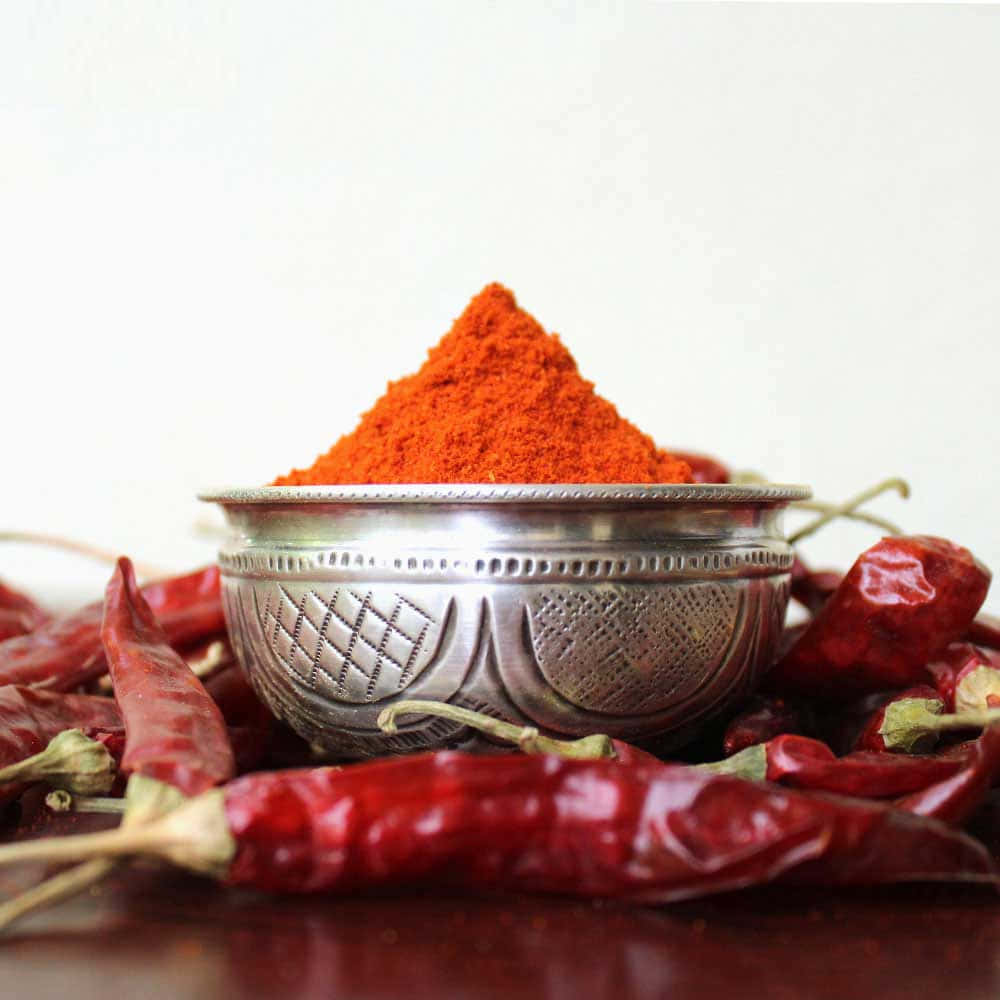 Red Chili Powder 桌布 壁紙 — 一個銀碗裝滿鮮豔的紅辣椒粉，周圍環繞著乾紅辣椒，放在深色表面上。