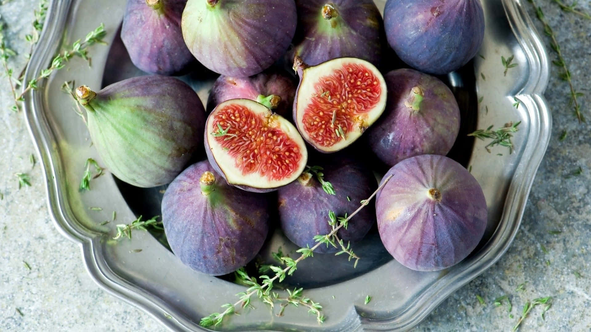 Purple Figs 桌布 壁紙 — 一個銀色扇貝形碟子盛載著成熟的紫色無花果，其中兩個切開露出粉紅色果肉，並有綠色百里香枝散落周圍。