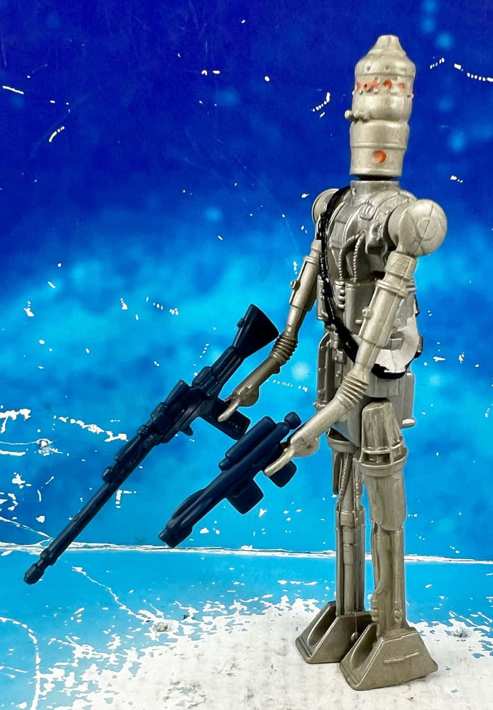 Ig-88 桌布 壁紙 — 一個銀色玩具機械人企喺度，喺有白色濺點嘅紋理藍色背景前，揸住兩把黑色爆能槍。
