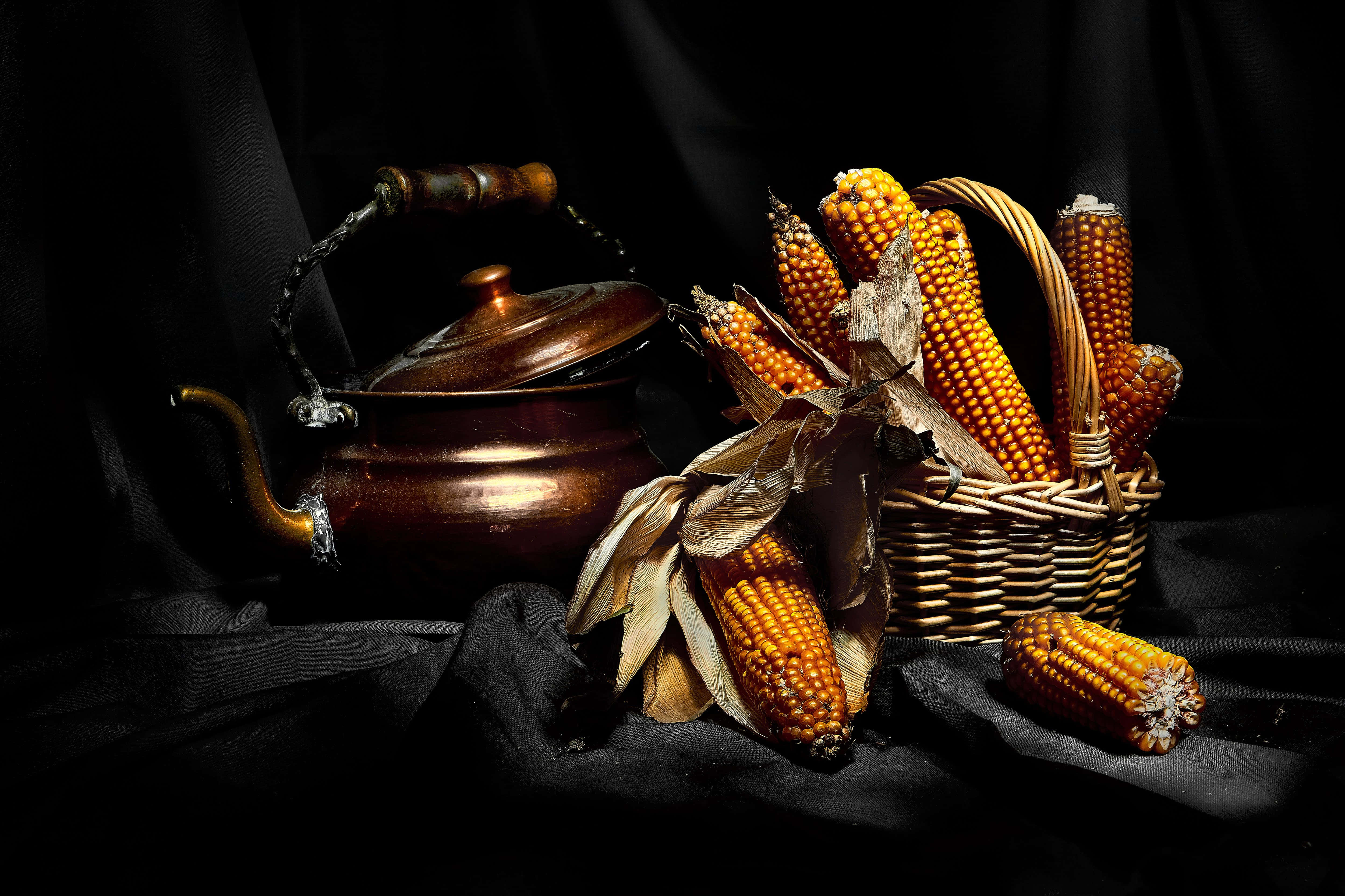 Corn 桌布 壁紙 — 一個銅壺和一籃金黃色的粟米放在深色布料上，在戲劇性的燈光下。