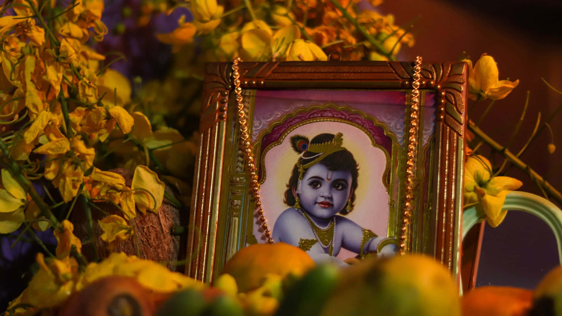Vishu 桌布 壁紙 — 一個鑲框的神聖兒童肖像，周圍環繞著鮮豔的黃色花朵和色彩繽紛的水果，在溫暖的燈光下。