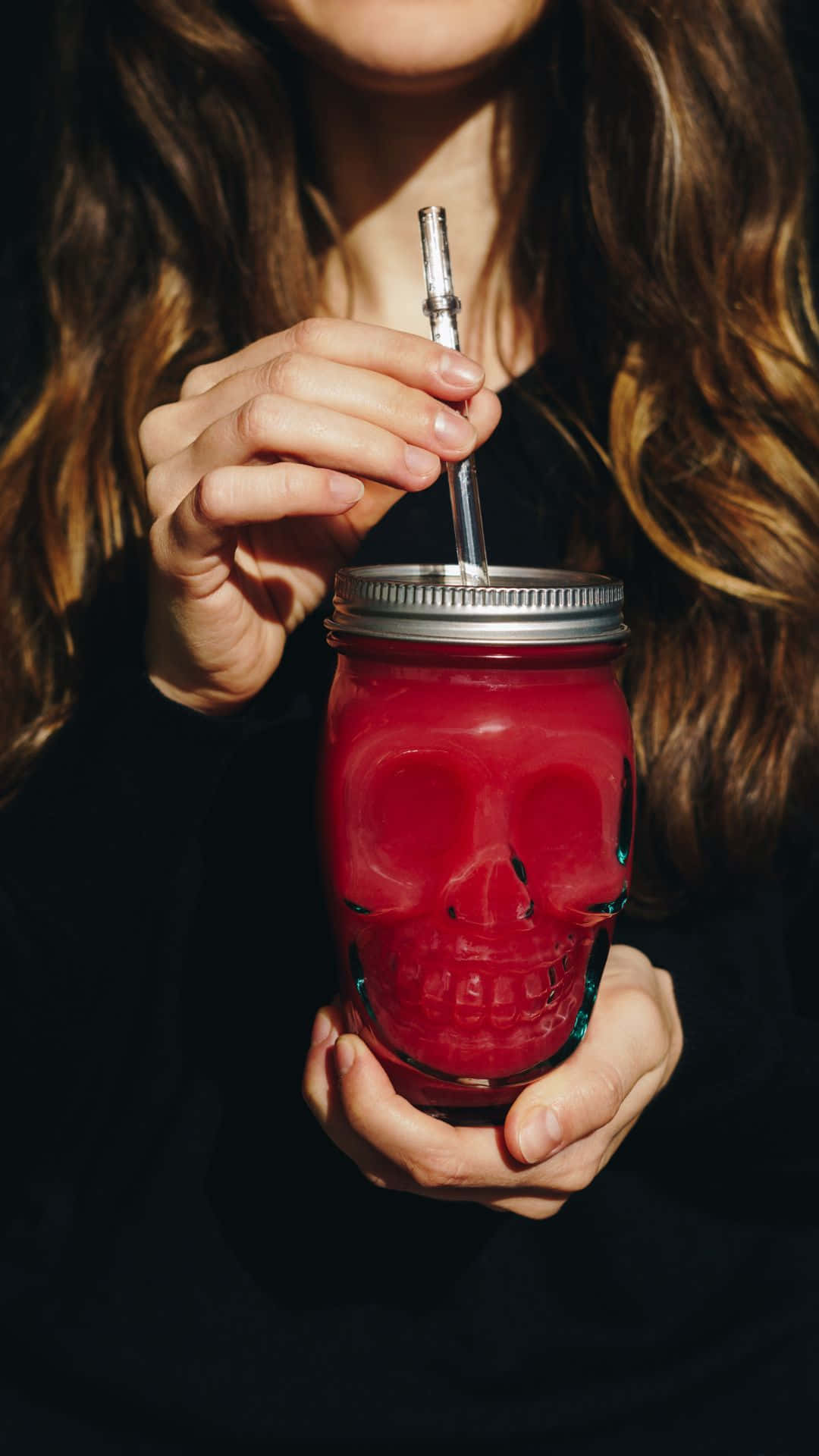 Spooky Cocktails 桌布 壁紙 — 一個長棕髮的人拿著一個裝有紅色液體、形狀像骷髏頭、帶有金屬吸管的玻璃瓶。