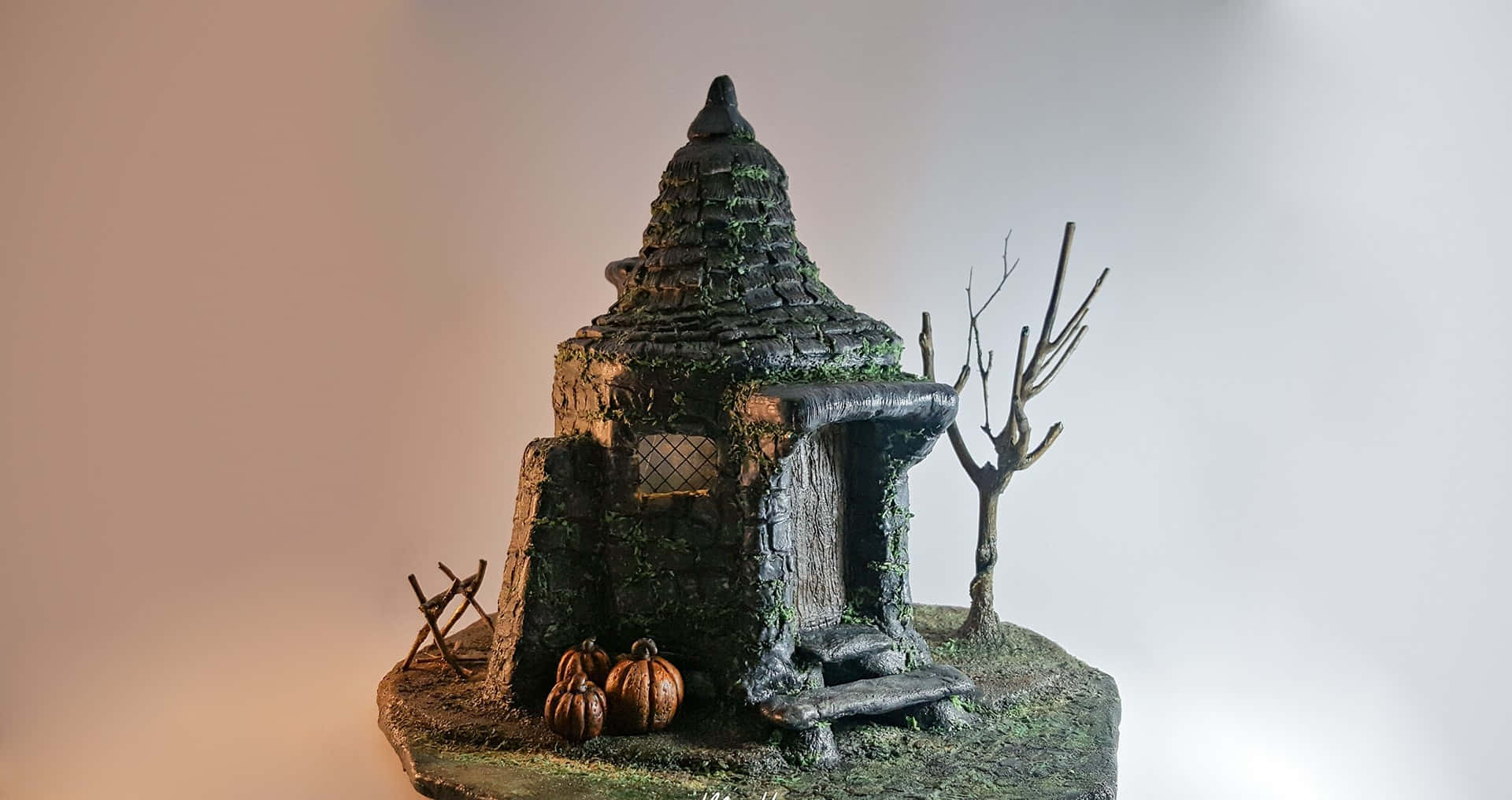 Hagrid's Hut 桌布 壁紙 — 一個長滿青苔的迷你鬼屋，放在岩石底座上，配有南瓜和一棵光禿禿的樹，背景是柔和的漸變色。