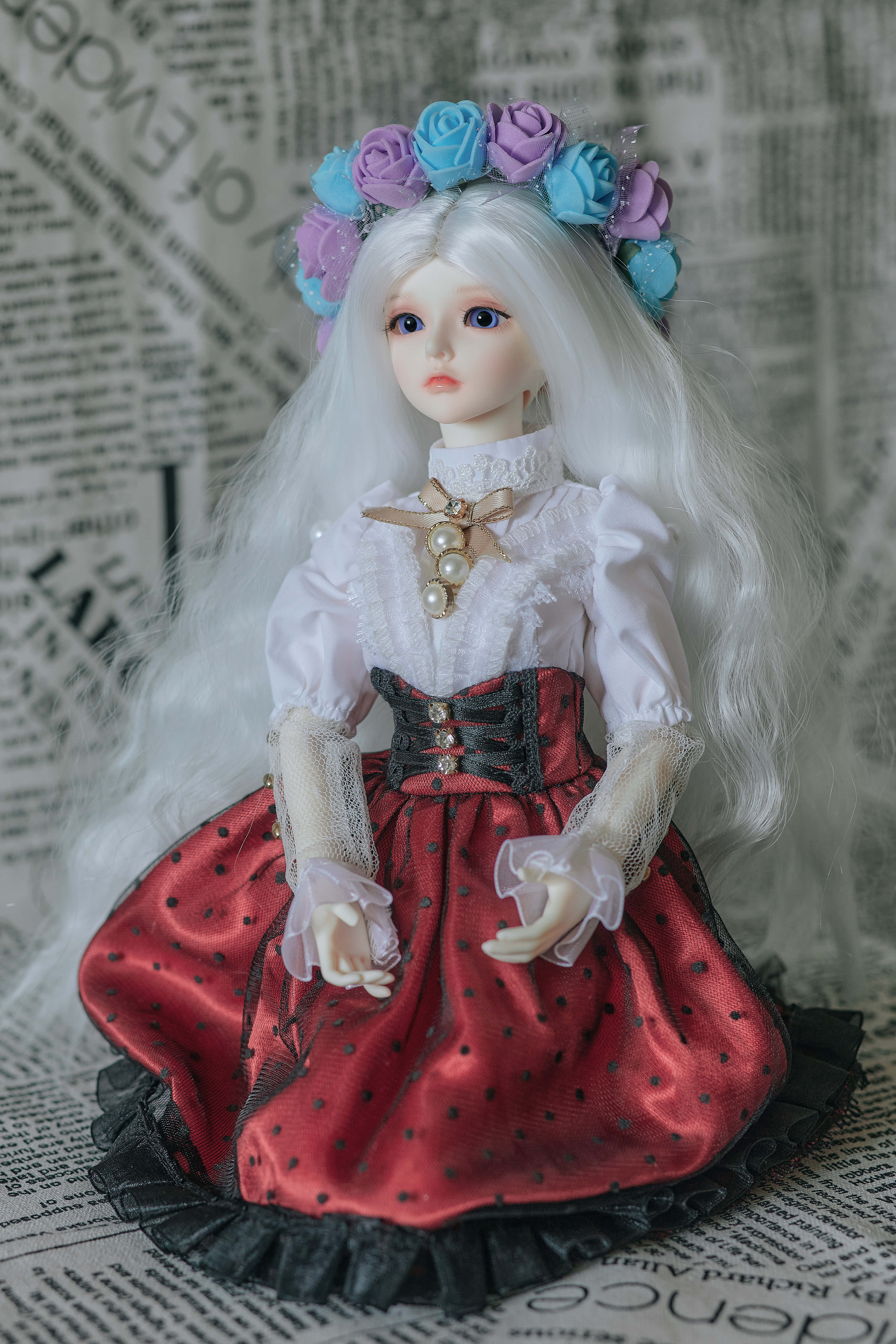 Dolls 桌布 壁紙 — 一個長白髮戴花冠的洋娃娃，穿著紅色圓點裙，優雅地坐在報紙背景前。