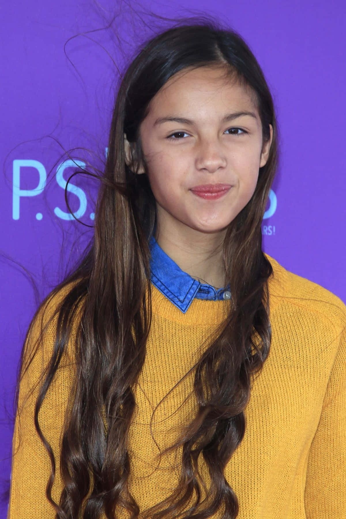 Olivia Rodrigo 桌布 壁紙 — 一個長著棕色長波浪髮的女孩，在紫色背景前穿著藍色有領襯衫外搭黃色毛衣，溫柔地微笑。