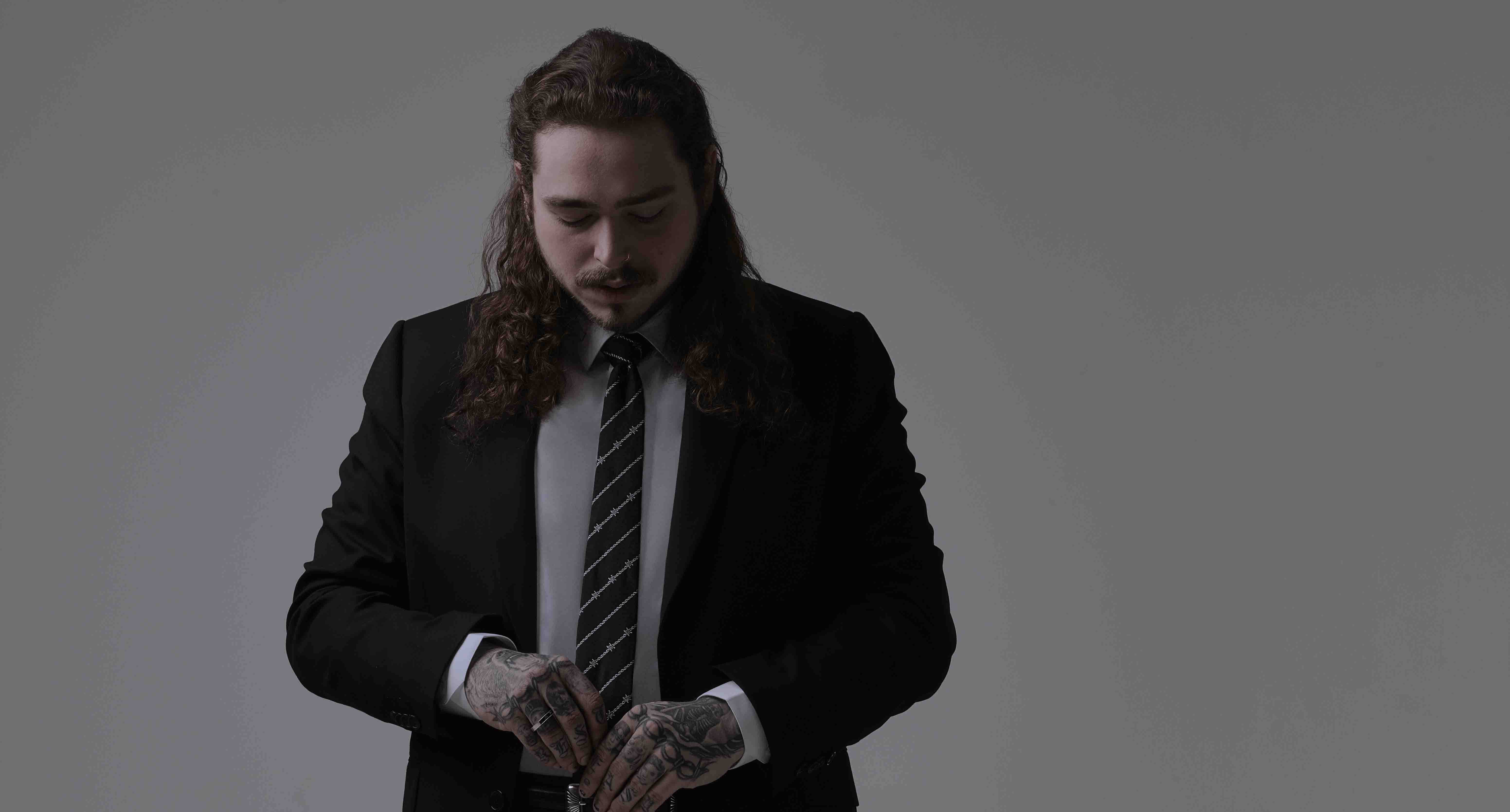 Post Malone Hits Magazine 2017 壁紙 — 一個長著長捲髮和紋身的男人在純灰色工作室背景前調整領帶。