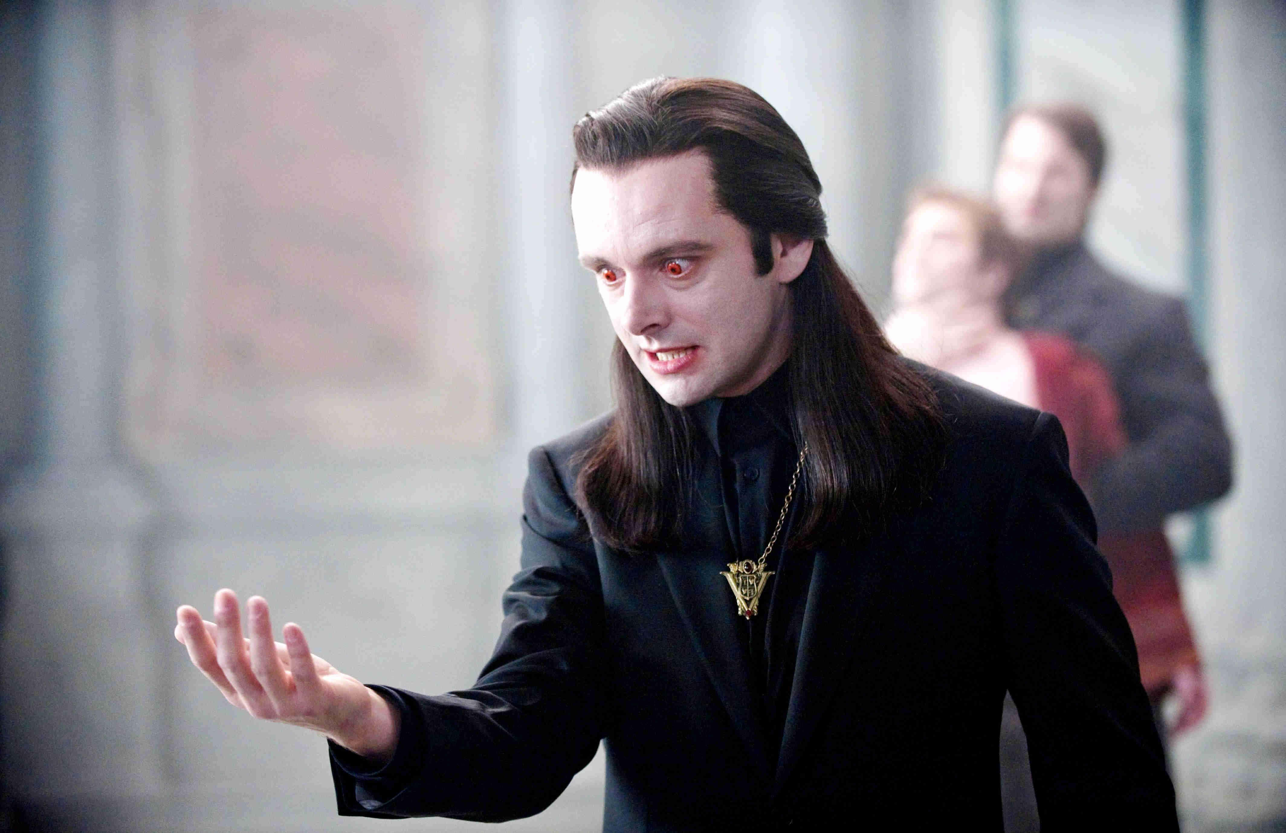 Michael Sheen 桌布 壁紙 — 一個長長黑髮、紅眼睛、身穿黑色西裝的吸血鬼，在昏暗的室內做出威脅性的手勢。