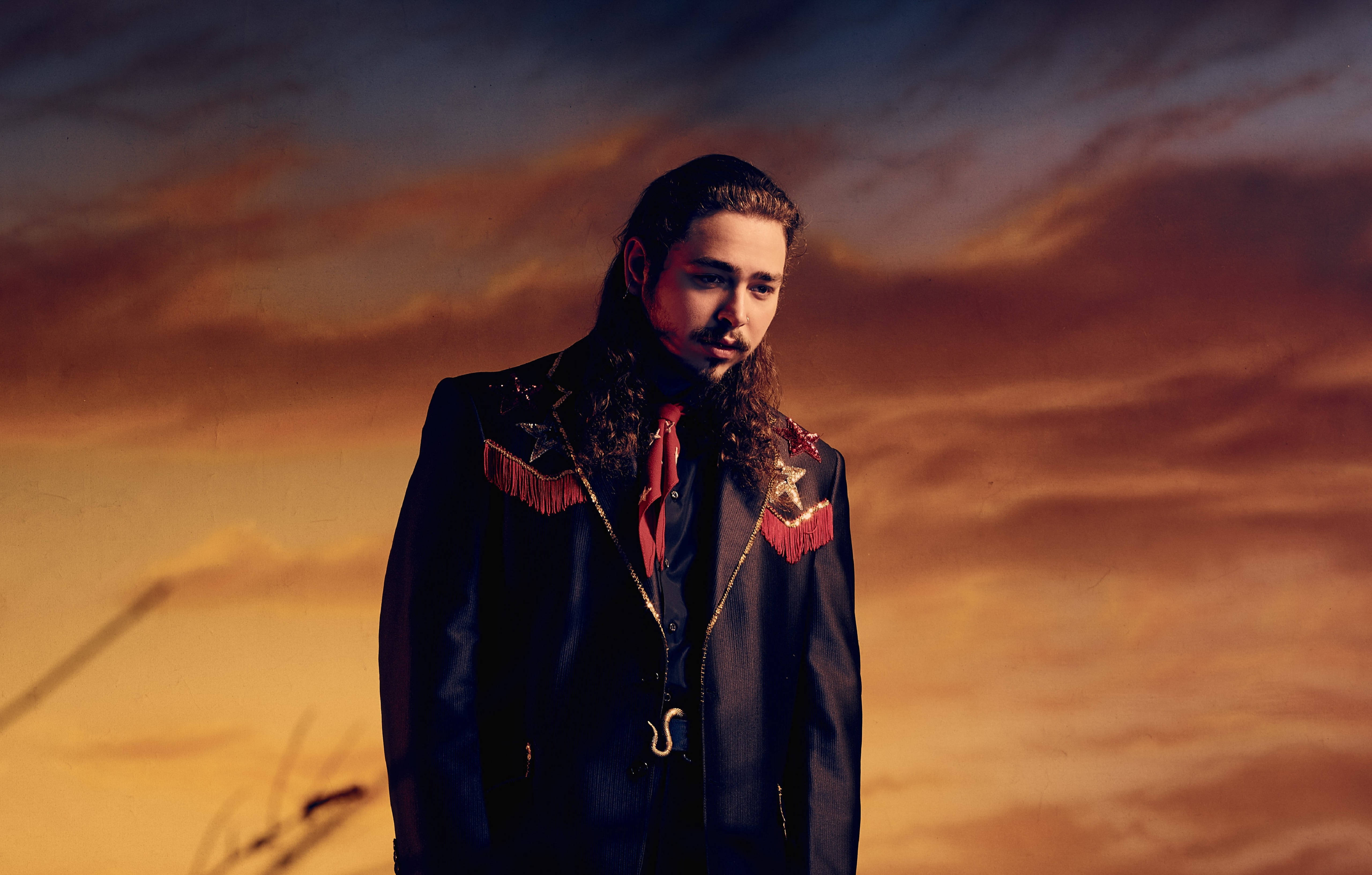 Post Malone Beerbongs Campaign 壁紙 — 一個長髮男子穿著流蘇黑色外套，站在戲劇性的日落天空前，天空有橙色和紫色的雲。