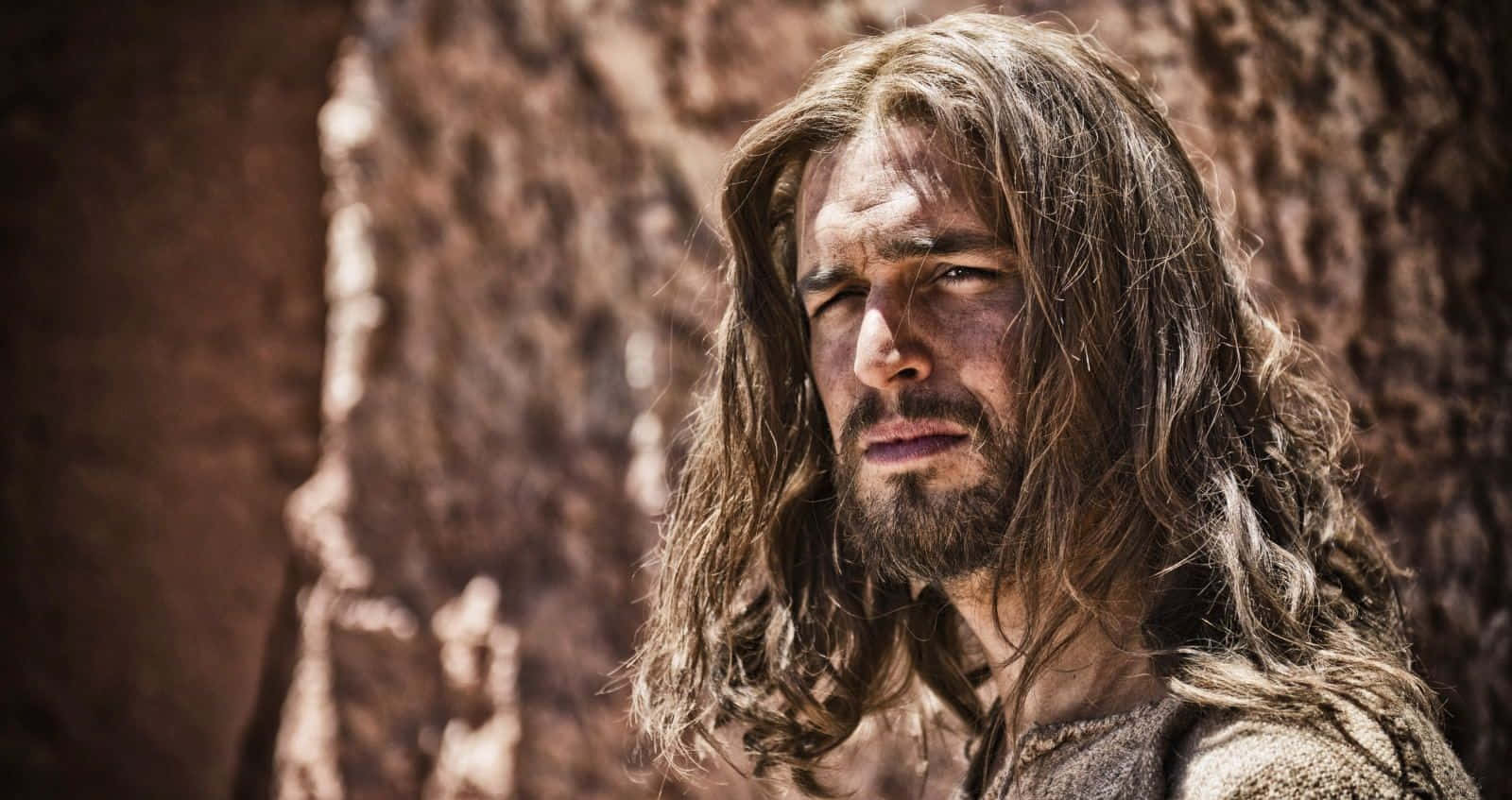 Son Of God 桌布 壁紙 — 一個長髮蓬鬆、留鬍子的男人在明亮的陽光下，對著崎嶇的岩石峭壁凝視。