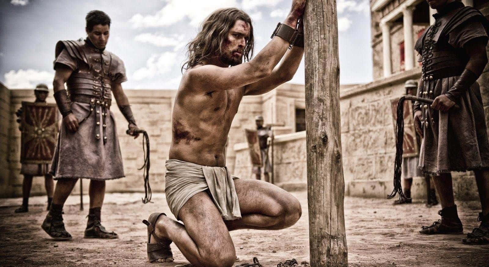 Son Of God 桌布 壁紙 — 一個長髮赤膊的男子在塵土飛揚的古老庭院中被羅馬士兵綁在木柱上。