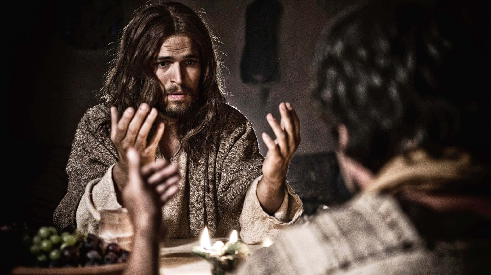 Son Of God 桌布 壁紙 — 一個長髮鬍鬚的男人坐在燭光桌旁，在與另一個人進行莊嚴的對話時做手勢。