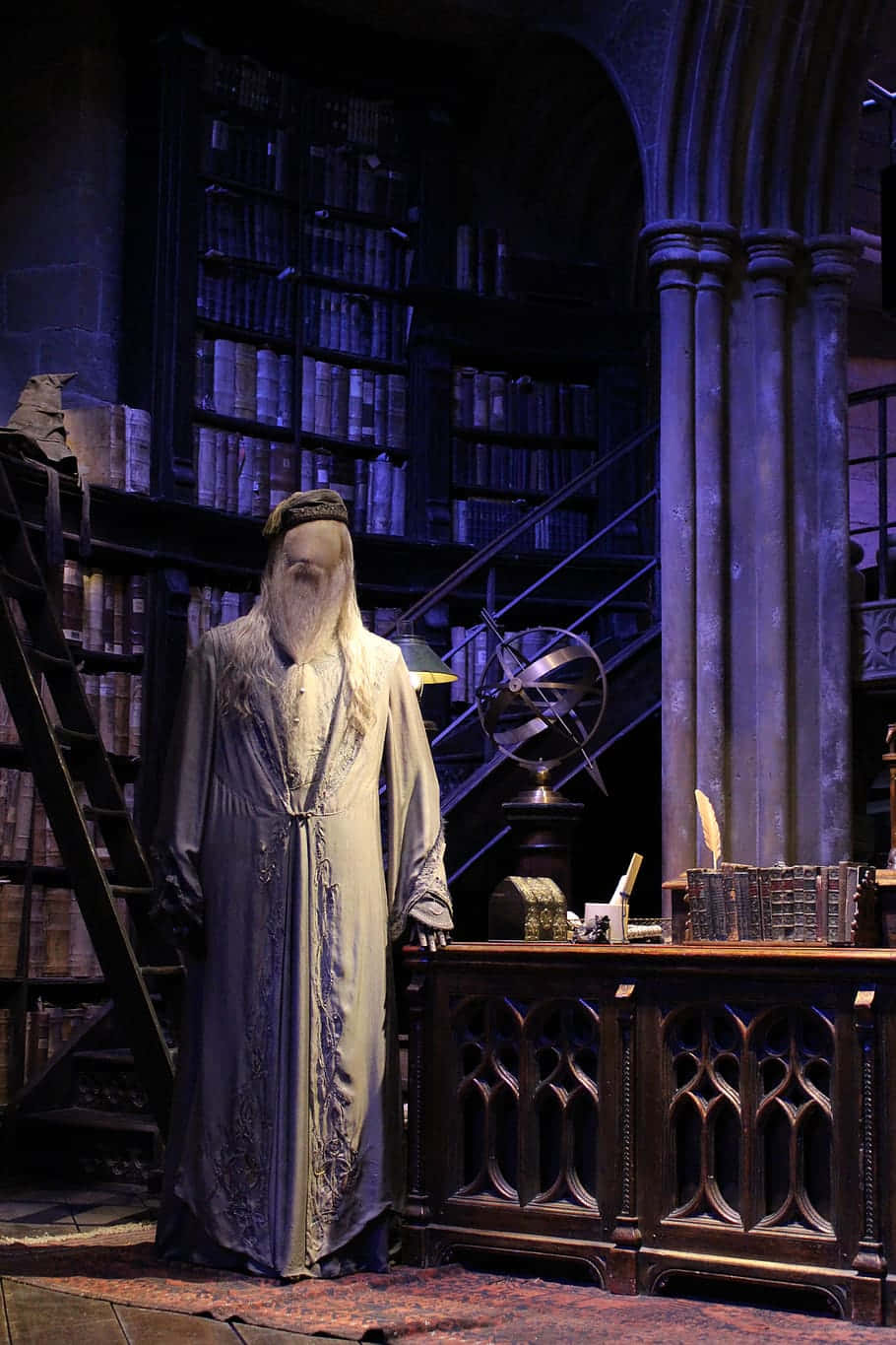 The Hogwarts Headmaster's Office 桌布 壁紙 — 一個長鬍鬚的幽靈巫師站在一個光線昏暗的哥德式圖書館中，圖書館內有高聳的書架和古董家具。