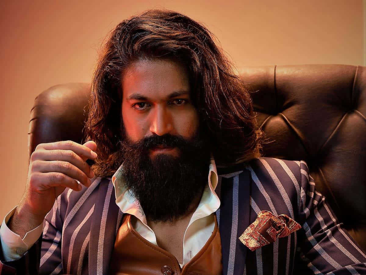 Kgf 2 桌布 壁紙 — 一個長黑髮、留滿鬍鬚的男人自信地坐在棕色皮椅上，身穿條紋西裝，手持香煙。