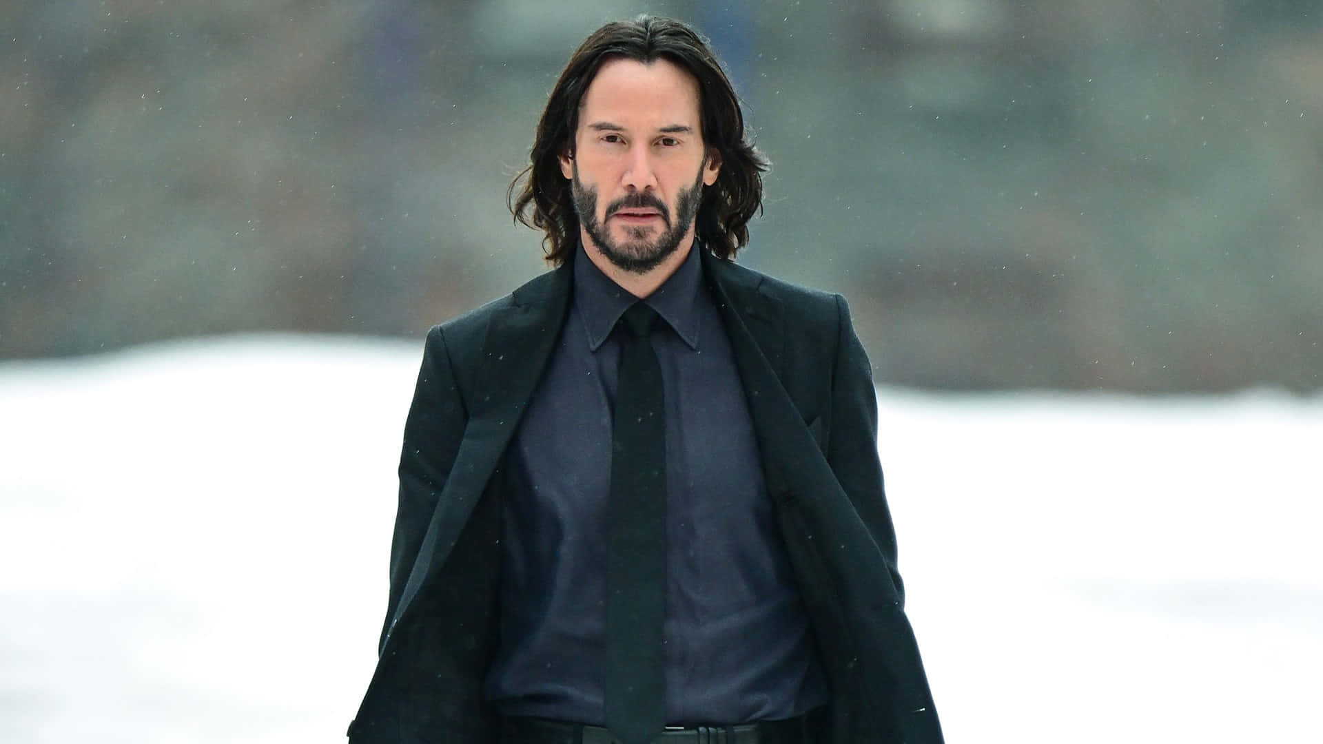 Keanu Reeves 桌布 壁紙 — 一個長黑髮、留鬍鬚、身穿黑色西裝的男人，在飄雪中莊嚴地站立，背景是模糊的冬日景色。