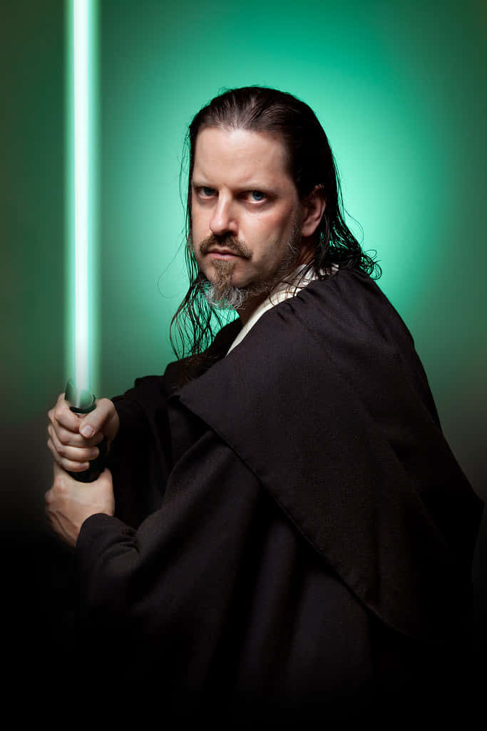 Qui-gon Jinn 桌布 壁紙 — 一個長黑髮、留鬍鬚、身穿黑色長袍的男人，在綠色漸變背景前手持發光的綠色光劍。