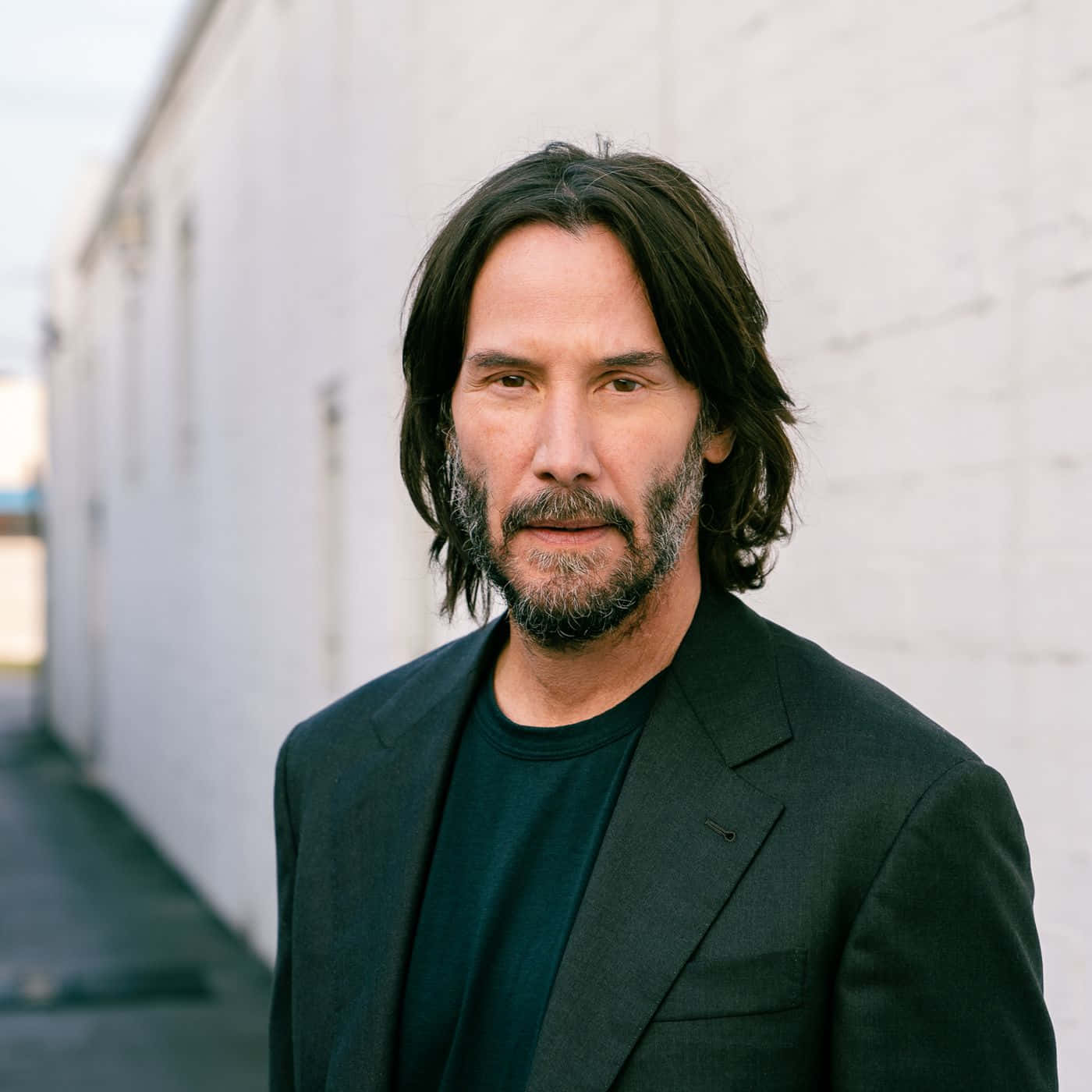 Keanu Reeves 桌布 壁紙 — 一個長黑髮留鬚的男人，身穿黑色西裝外套內搭黑色T恤，站在白色磚牆前。