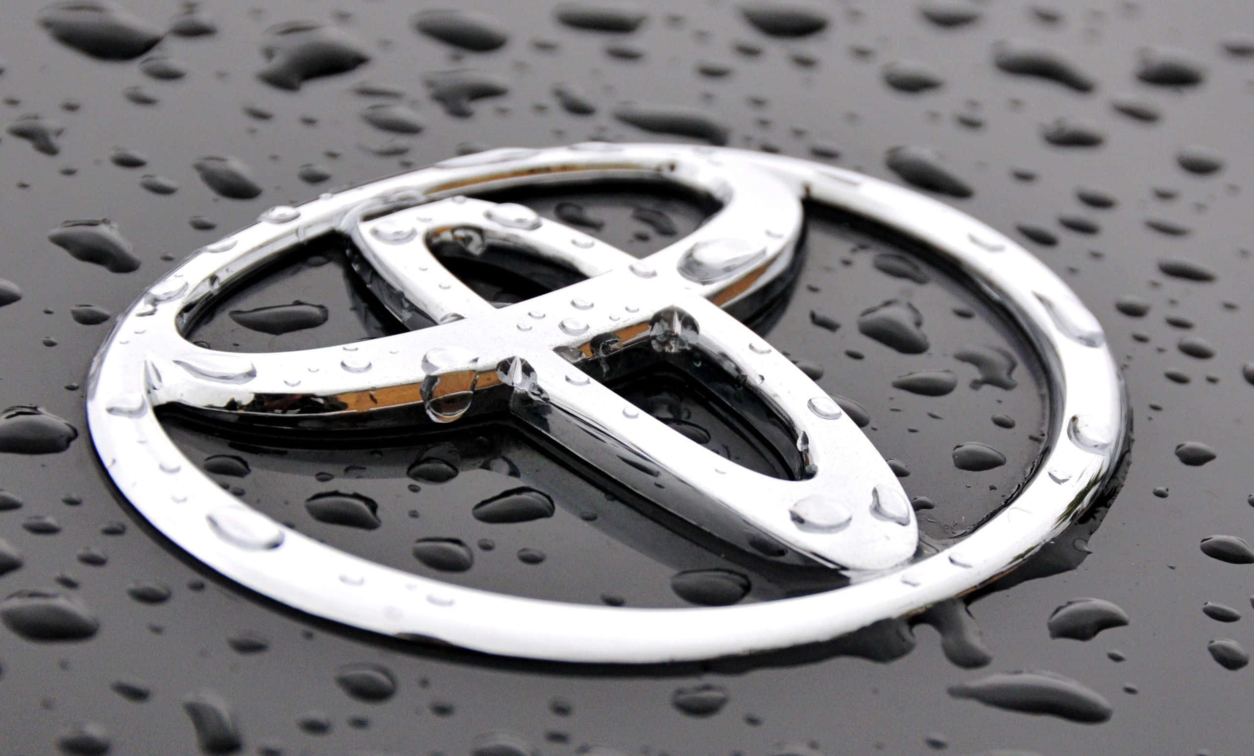 Toyota 桌布 壁紙 — 一個閃亮嘅鍍鉻豐田標誌近距離特寫，喺濕嘅黑色引擎蓋上，雨滴閃閃發光。