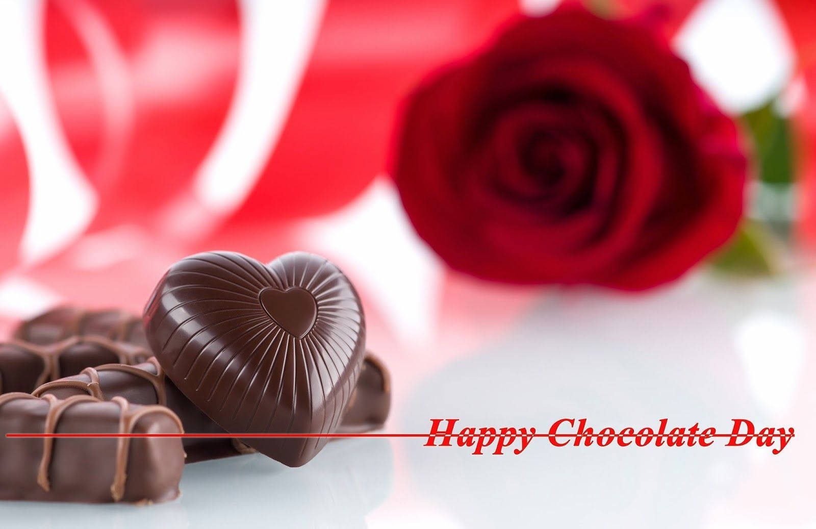 Chocolate Day 桌布 壁紙 — 一個閃亮的心形朱古力放在一朵鮮紅玫瑰旁邊，背景是模糊的紅色和白色，並有「Happy Chocolate Day」文字。