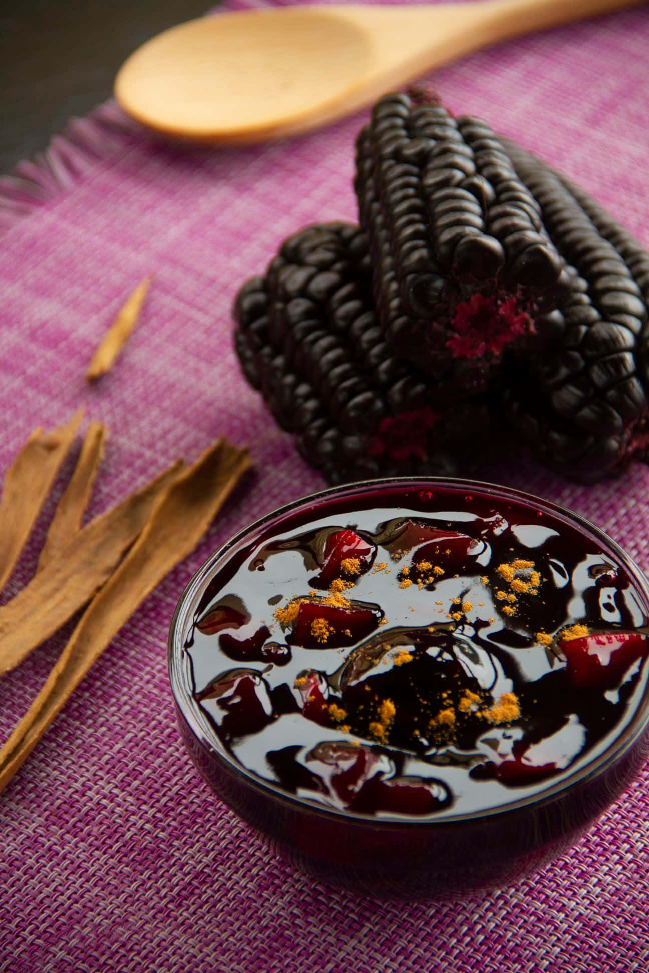 Purple Corn 桌布 壁紙 — 一個閃亮的深色雜莓蜜餞碗，上面撒有肉桂粉，旁邊放著新鮮黑莓和肉桂棒，置於有紋理的紫色布上。