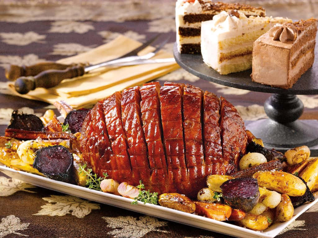 Black Forest Ham 桌布 壁紙 — 一個閃亮的烤火腿，切好的片放在大盤上，周圍是烤蔬菜，並用新鮮香草裝飾。