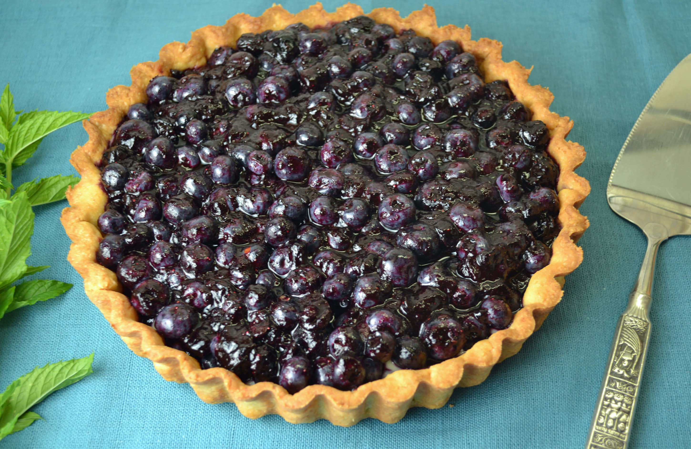 Blueberries Tart 桌布 壁紙 — 一個閃亮的藍莓撻，金黃色的撻皮放在青綠色布料上，旁邊有薄荷葉和裝飾用服務鏟。