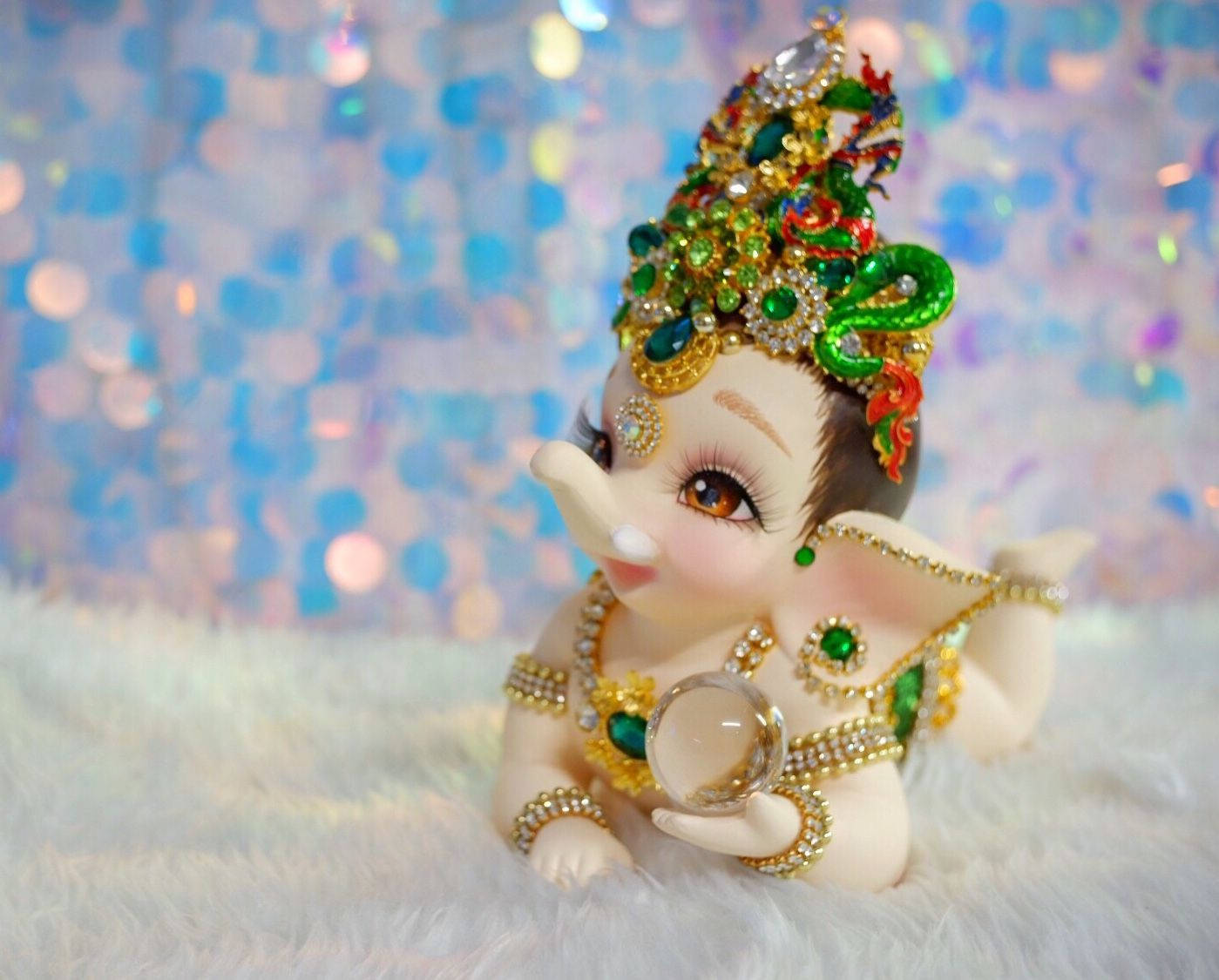 Baby Ganesh 桌布 壁紙 — 一個閃閃發光的小象雕像，飾有珠寶和皇冠，坐在蓬鬆的白色毛皮上，背景是閃爍的藍色。