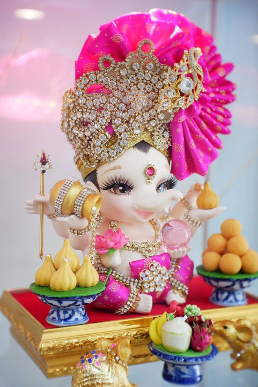 Baby Ganesh 桌布 壁紙 — 一個閃閃發光的粉紅色和金色嬰兒神像，飾有珠寶，在華麗的金色基座上拿著糖果。