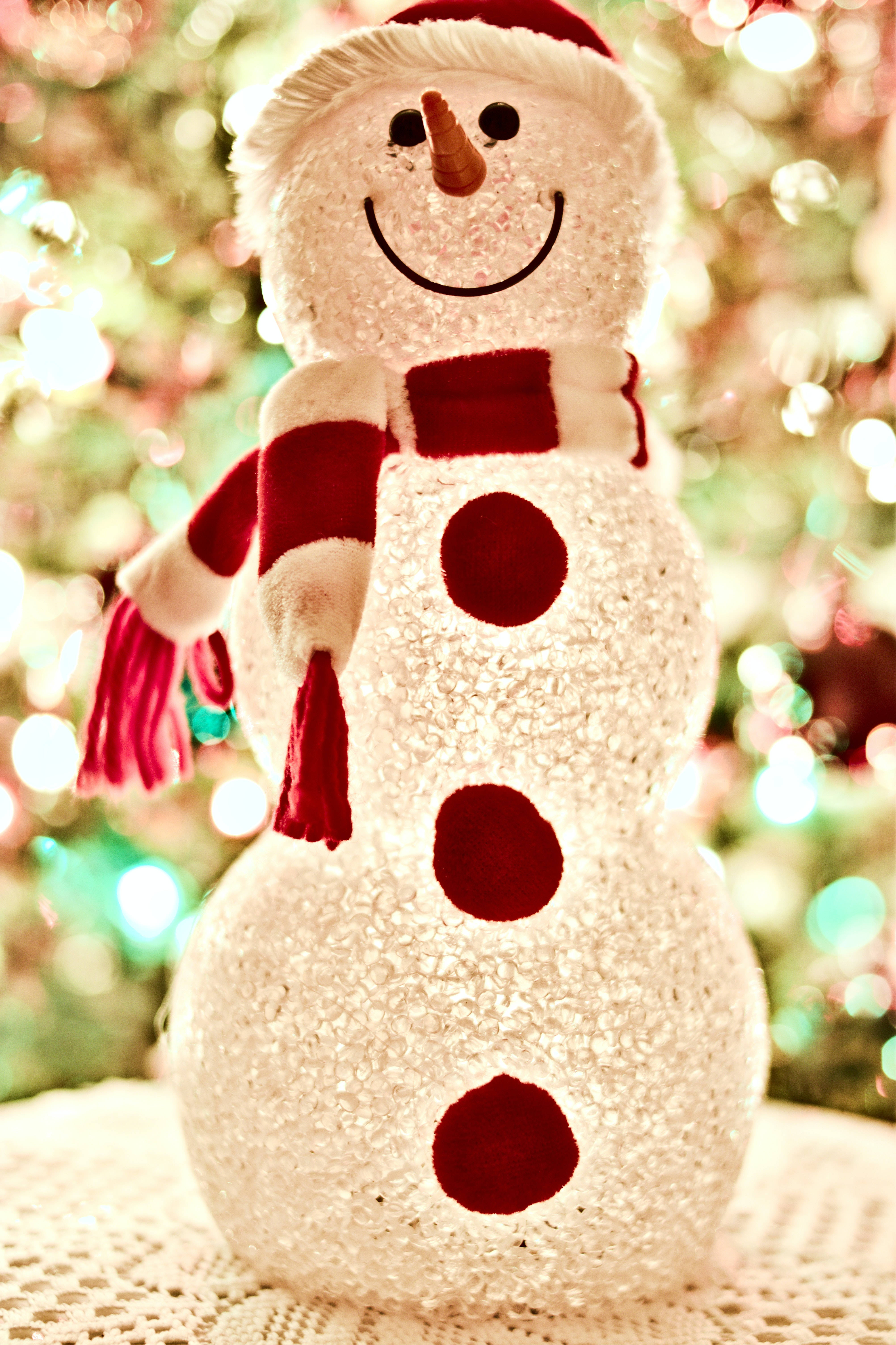 雪人 桌布 壁紙 — 一個閃閃發光的雪人公仔，戴著紅色圍巾和帽子，在散景聖誕樹背景下溫暖地微笑。