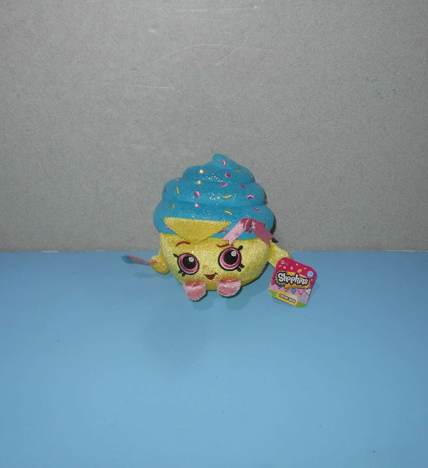 Shopkins 桌布 壁紙 — 一個閃閃發光的黃色杯子蛋糕毛絨玩具，帶有藍色糖霜和粉紅色糖粒，放在藍色表面上，背景是灰色牆壁。