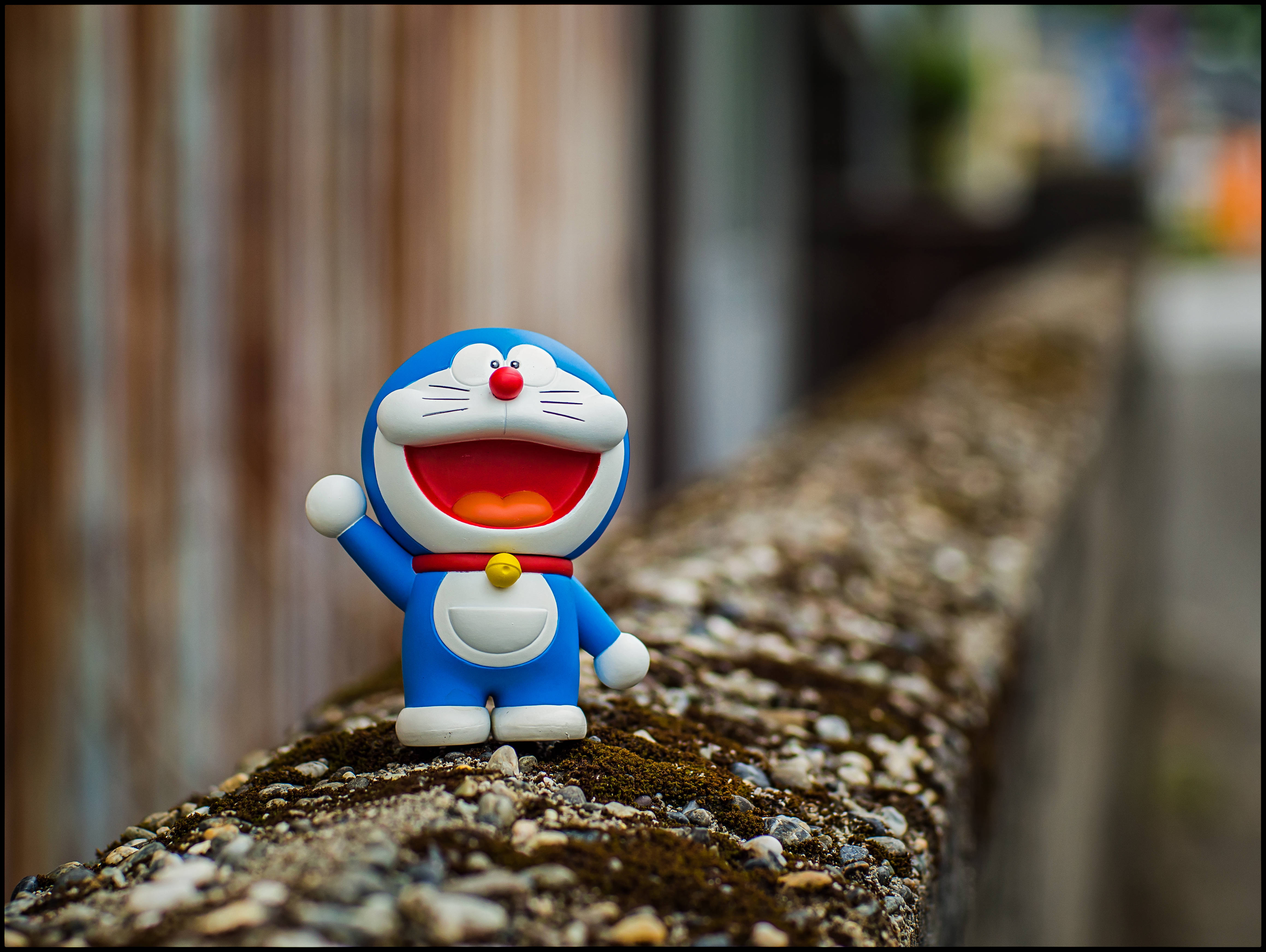 Doraemon 桌布 壁紙 — 一個開心嘅藍色多啦A夢公仔，張大個嘴，舉起一隻手，企喺一個生咗苔蘚嘅石壆上面，背景係模糊嘅木紋。