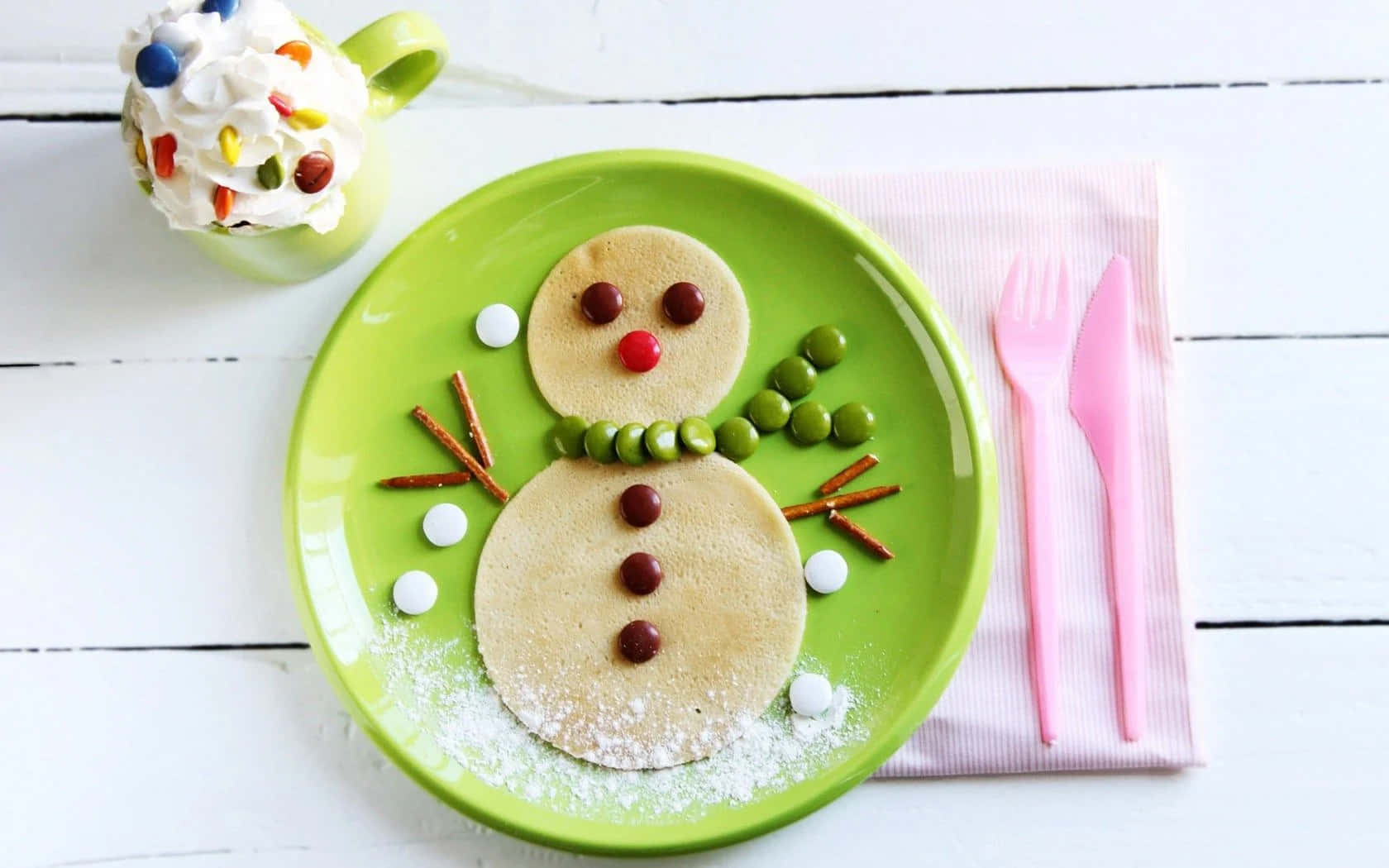 Winter Food 桌布 壁紙 — 一個開心雪人煎餅放在綠色碟上，有糖果眼睛、椒鹽卷餅手臂和糖粉雪，旁邊有節日杯和粉紅色餐具。
