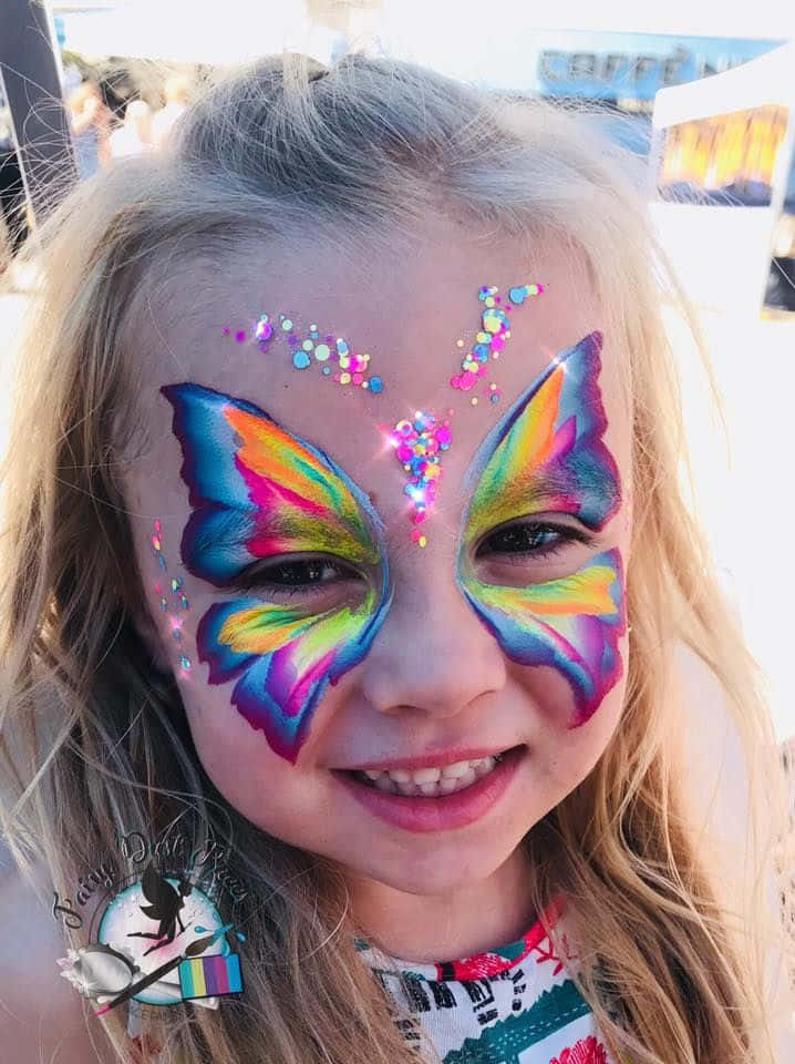 Butterfly Face Painting 桌布 壁紙 — 一個開朗的小女孩，臉上畫著鮮豔的彩虹蝴蝶彩繪和閃粉，對著鏡頭燦爛地微笑。