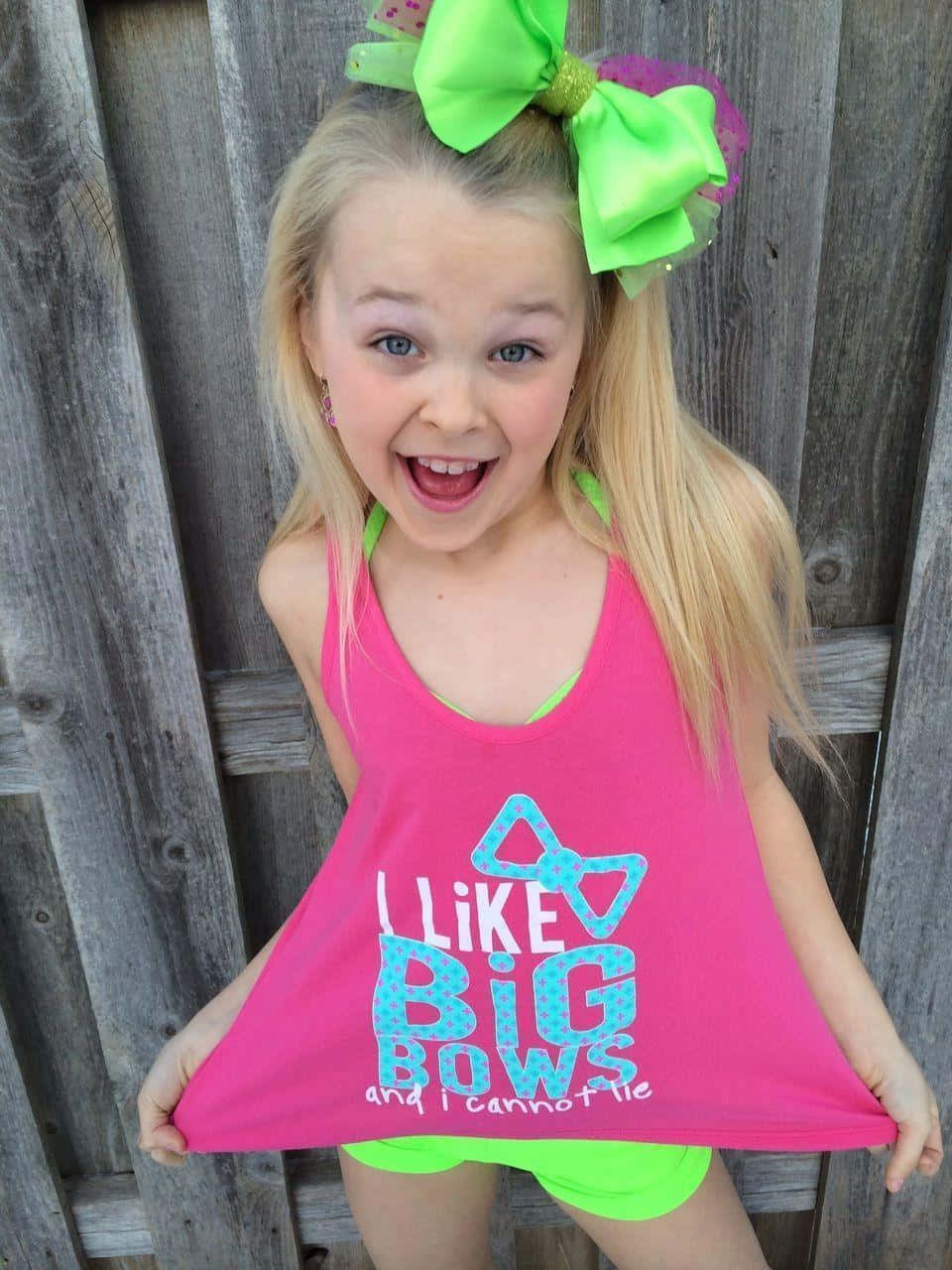 jojo siwa 桌布 壁紙 — 一個開朗的金髮小女孩，戴著一個巨大的螢光綠色蝴蝶結，穿著粉紅色背心和短褲，對著風化的木柵欄咧嘴大笑。