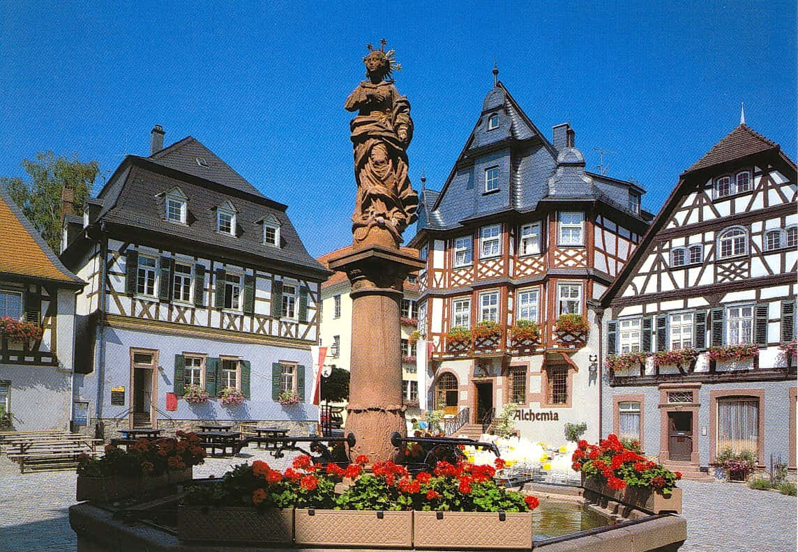 Germany 桌布 壁紙 — 一個陽光明媚的歐洲小鎮廣場，中央有一座石像噴泉，周圍是半木結構建築和鮮豔的紅色花箱。