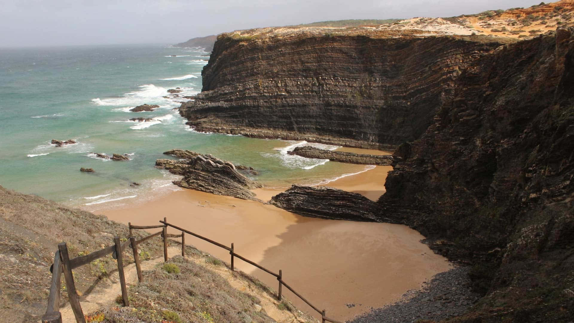 Portuguese Beach 桌布 壁紙 — 一個隱蔽的小海灣，金色沙灘 nestled 在高聳的層狀懸崖之間，海浪拍打岩石。