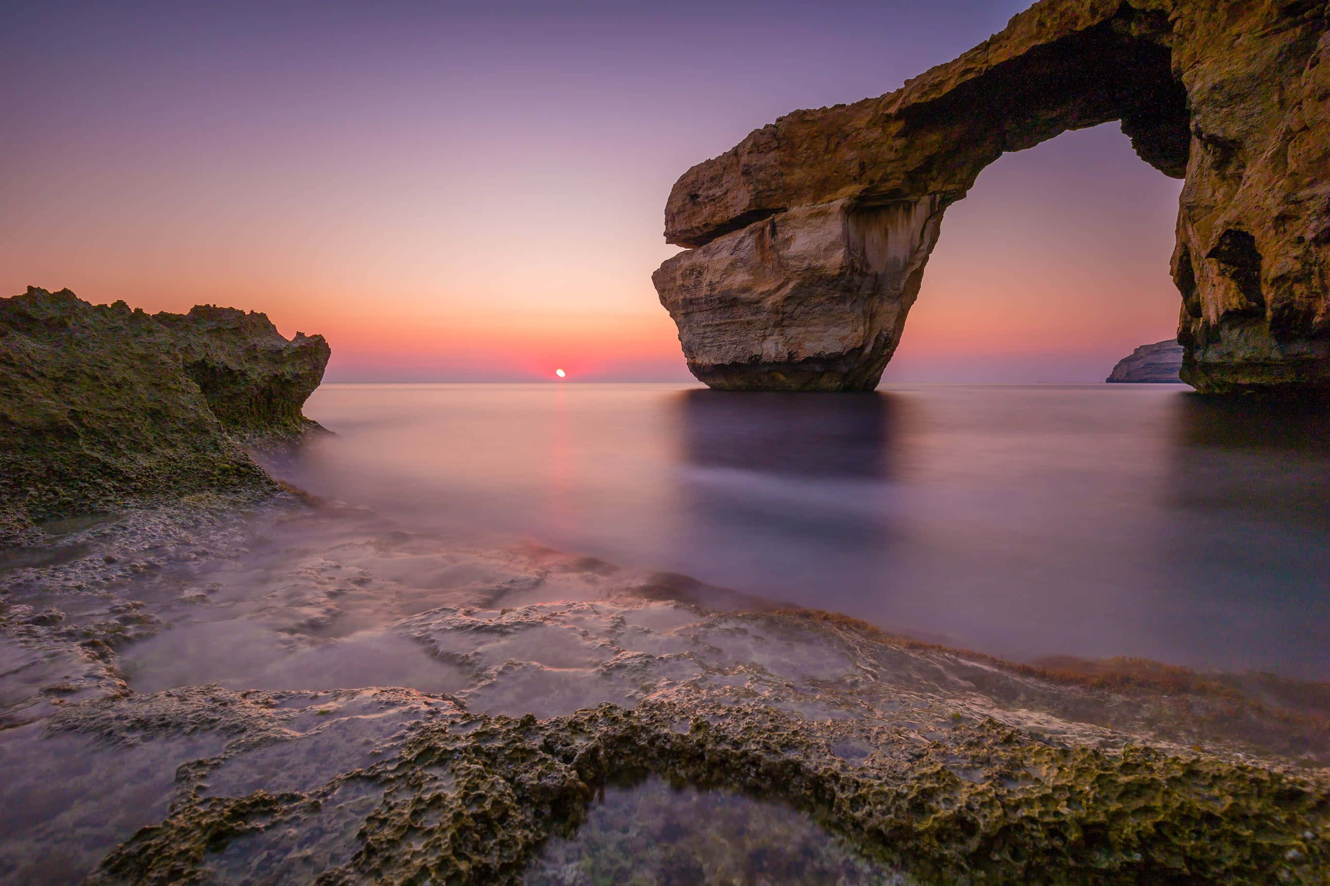 Malta 桌布 壁紙 — 一個雄偉的自然岩石拱門在日落時發光，下方有平靜的水流，上方是充滿活力的紫橙色天空。