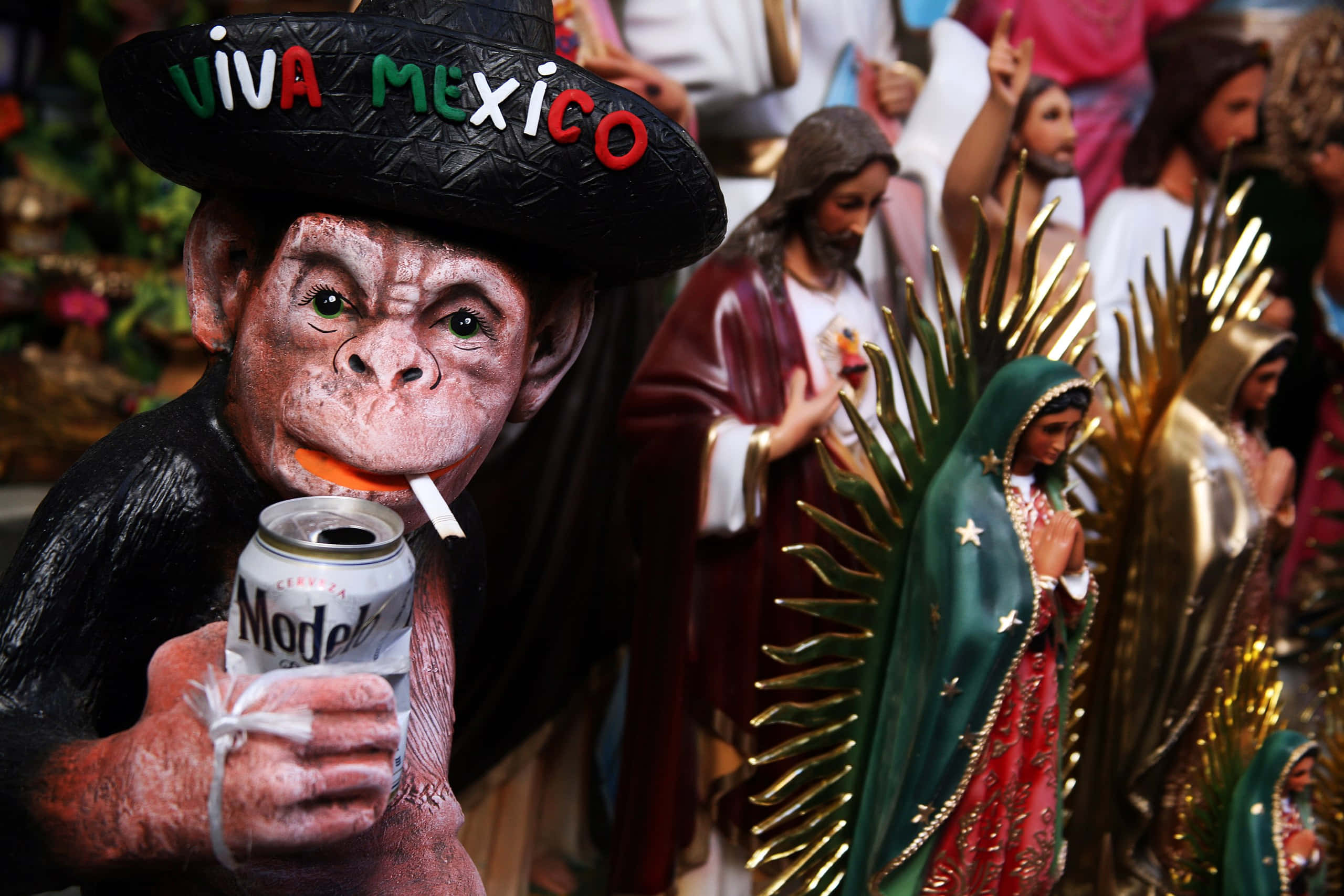 墨西哥 桌布 壁紙 — 一個雕刻的猴子公仔戴住「Viva Mexico」闊邊帽，喺宗教雕像旁邊攞住一罐Modelo同埋一支煙。