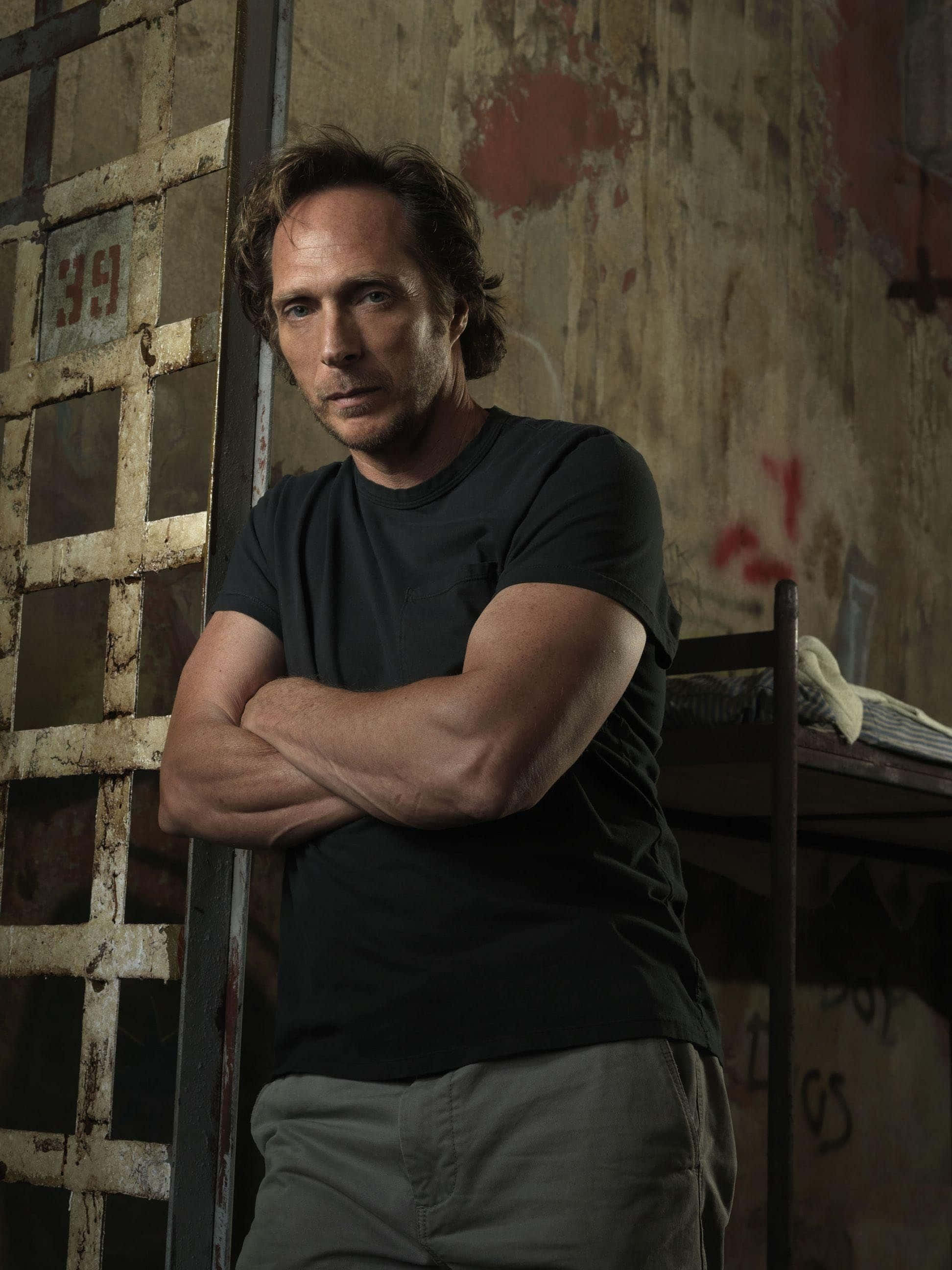 William Fichtner 桌布 壁紙 — 一個雙臂交叉的男人站在一個粗糙的監獄牢房裡，生鏽的鐵欄和剝落的牆壁。